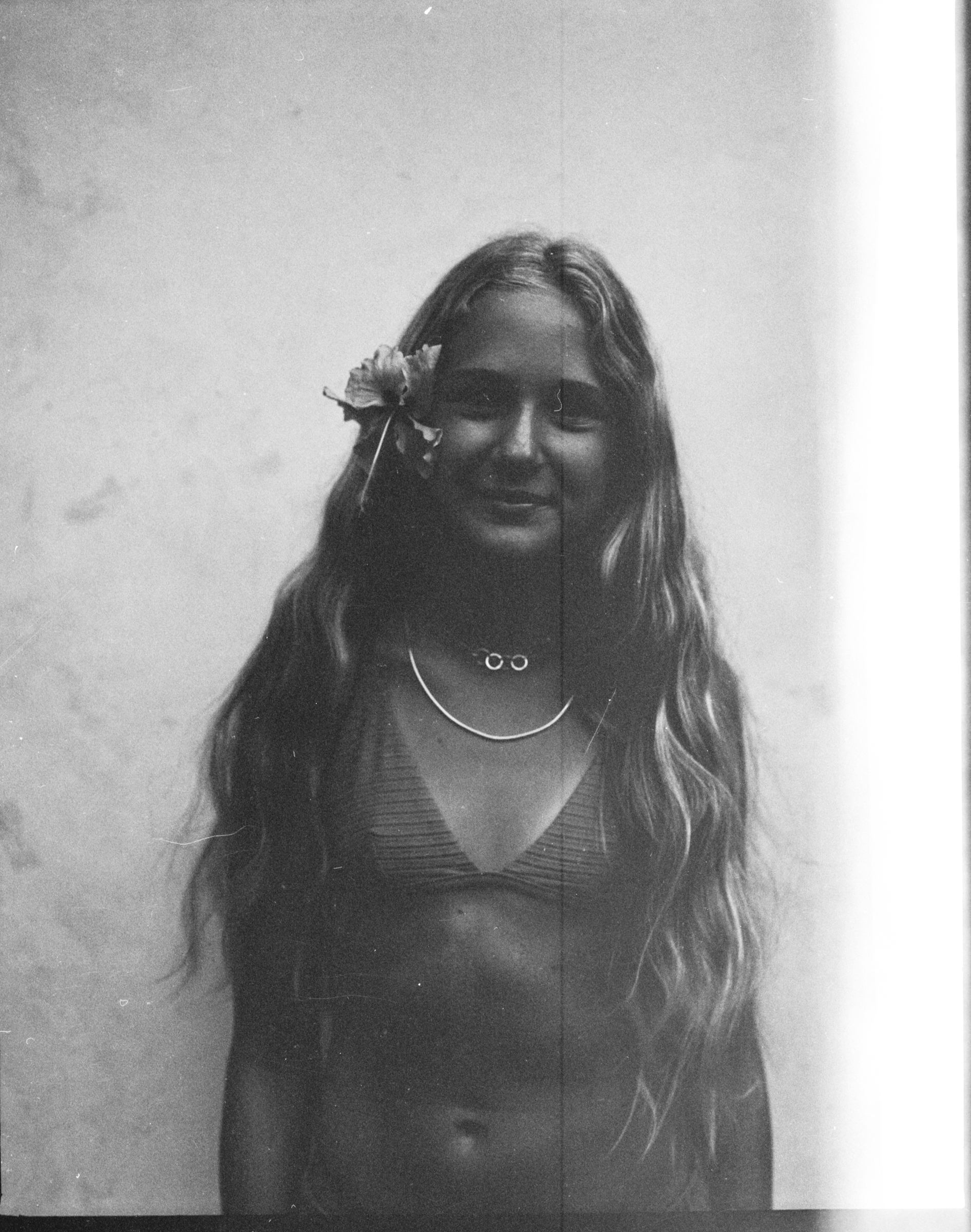 Black and white photograph of a woman with long wavy hair, a flower in hair, wearing necklaces and a striped top, standing against a plain wall.