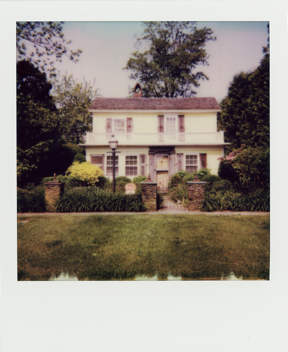A two-story house with a porch, surrounded by a green lawn and trees, with a lamp post in front.