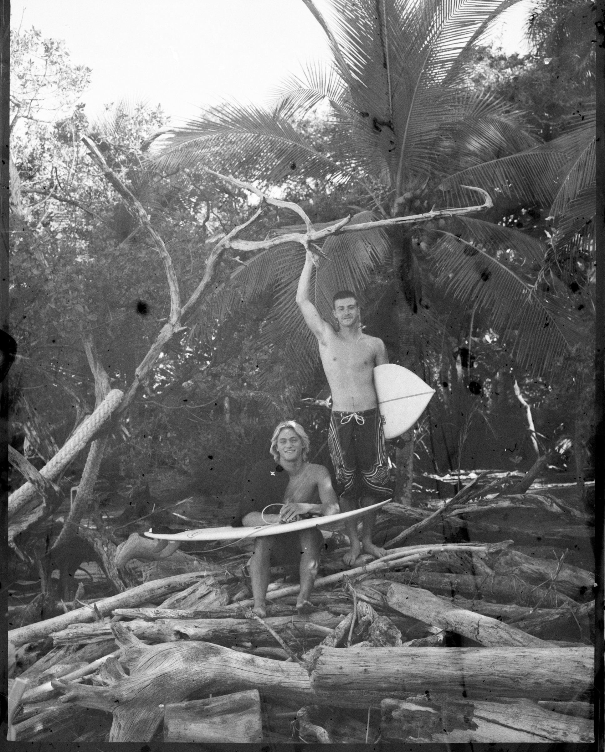 Two young men with surfboards, one shirtless, smiling, standing and sitting on logs amid tropical trees, in black and white.