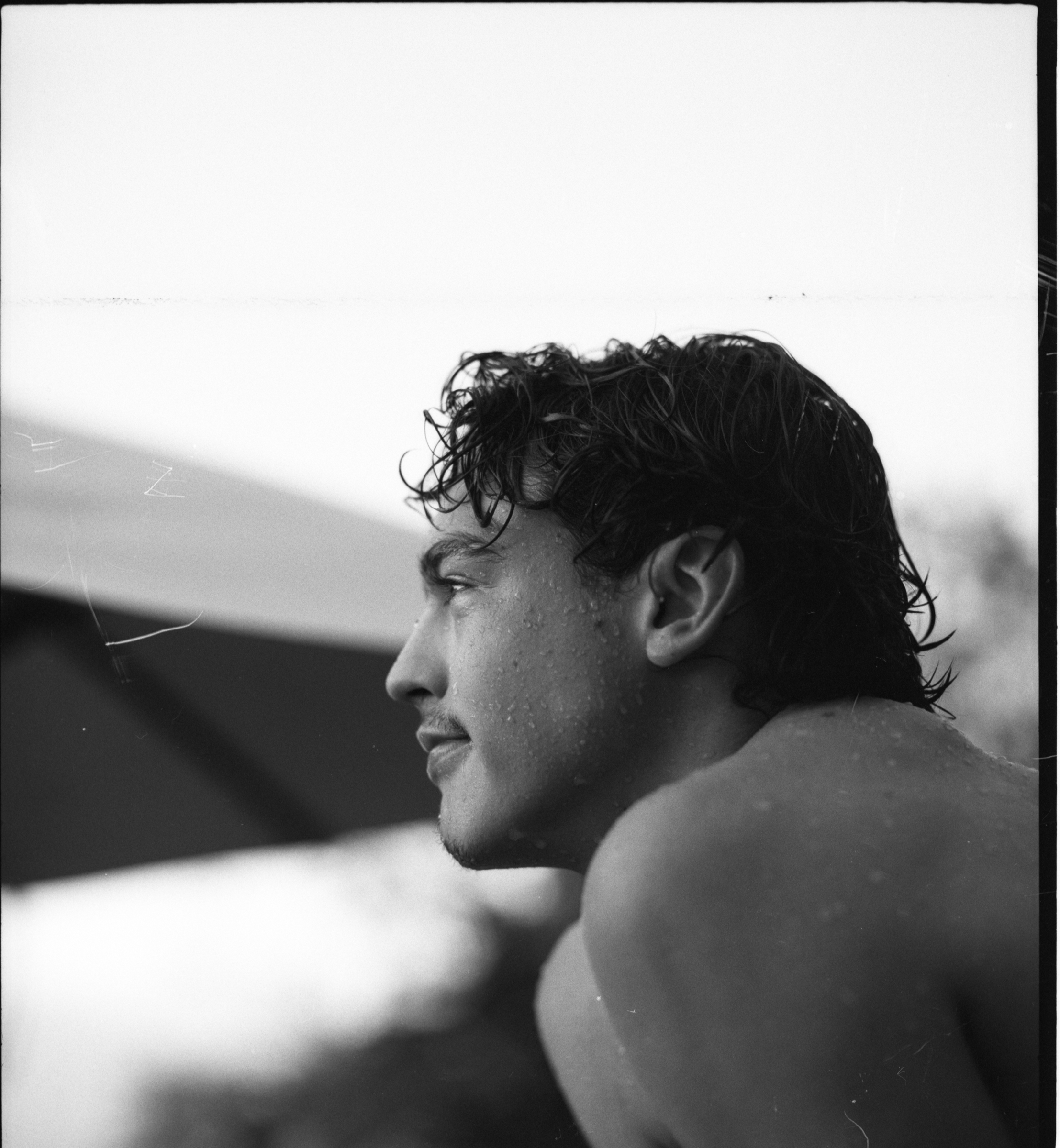 Close-up black and white photo of a young man with wet, curly hair, looking to the left, with water droplets on his face and shoulder.