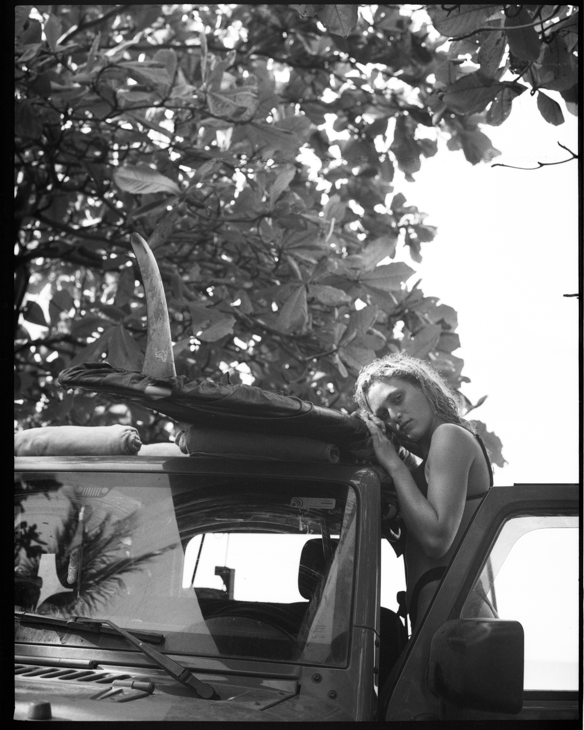 A woman with curly hair leans on the side of a parked vehicle, with a surfboard and rolled-up towel on top of the vehicle. She appears to be resting or looking contemplatively, with lush foliage in the background.