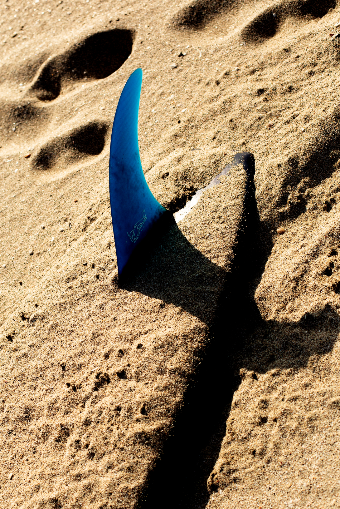Blue surfboard, True Ames single fin planted in sand at the shoreline, surf lifestyle detail shot