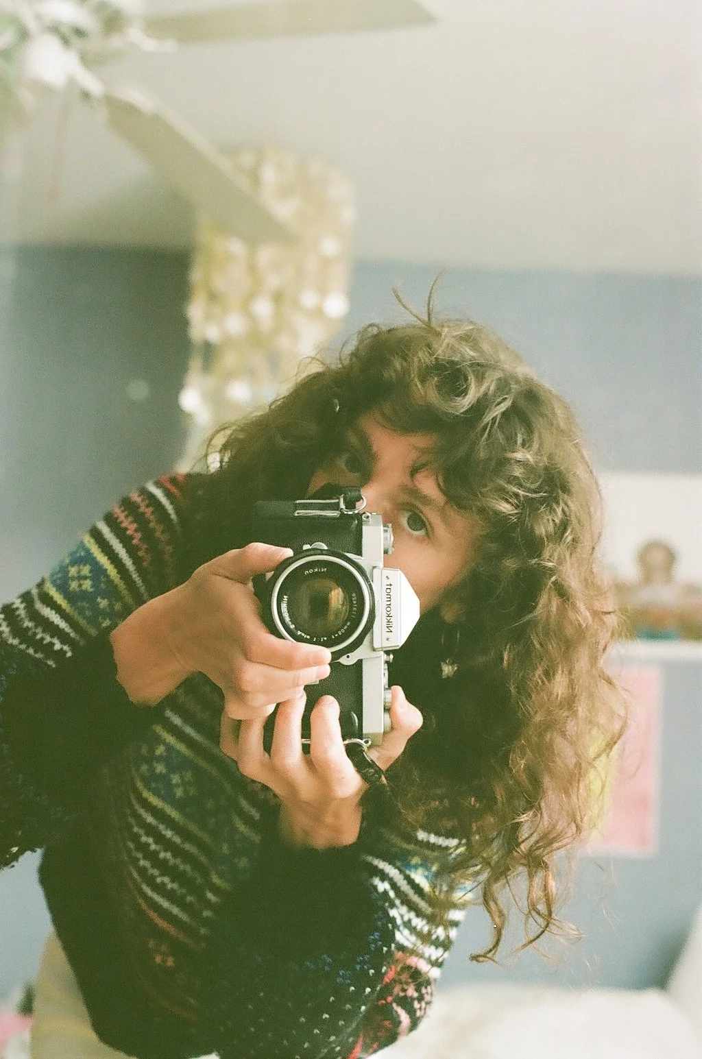 A person with curly hair taking a photo of themselves with a vintage camera in a mirror.