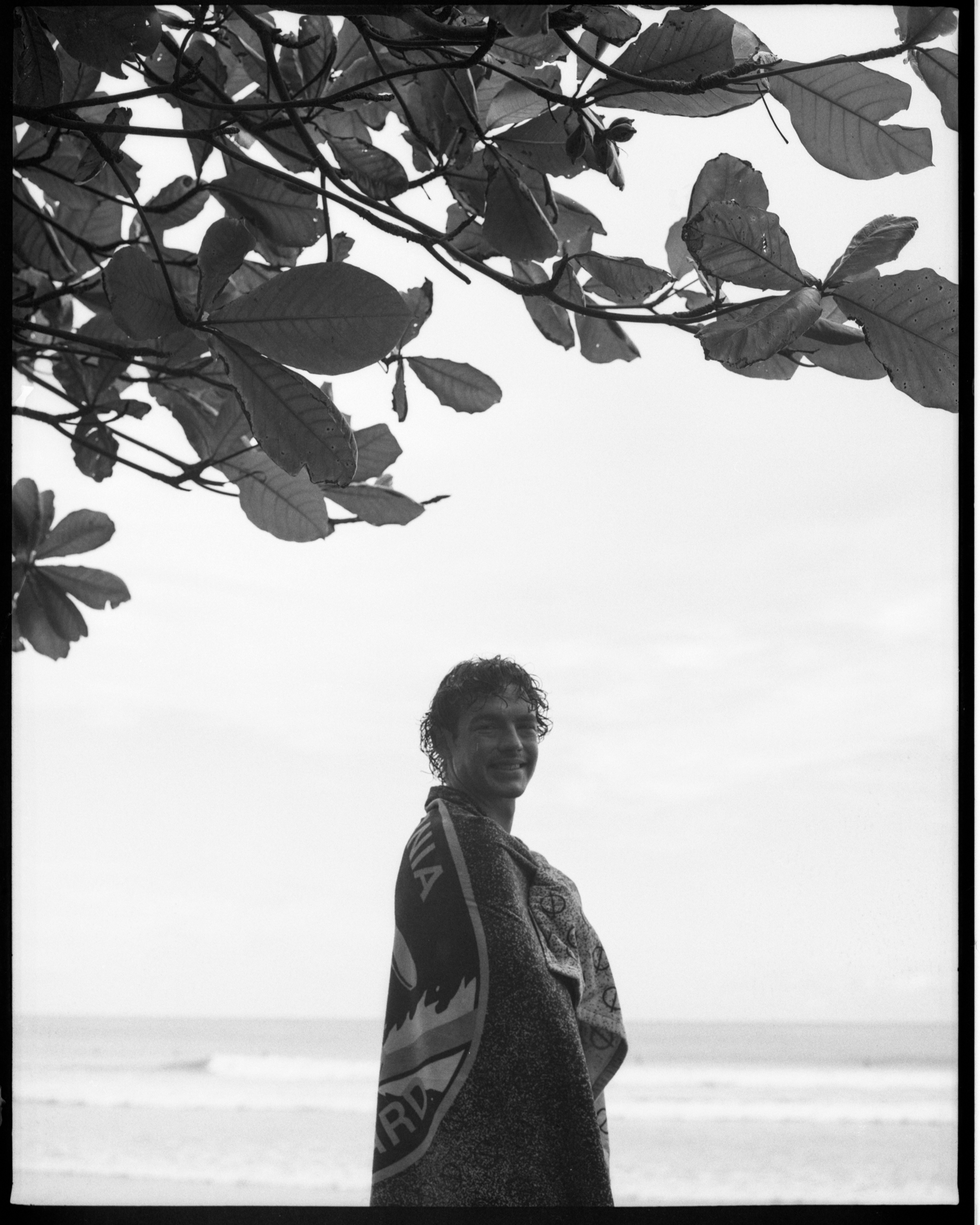 A young man smiling on the beach, wrapped in a patterned towel, with the ocean in the background and overhanging tree branches in the foreground.