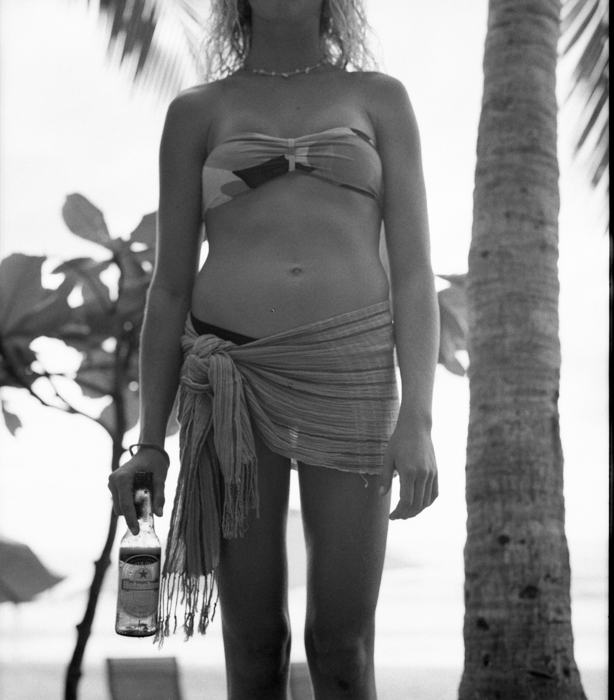A woman standing outdoors near a palm tree, wearing a strapless top and a sarong. She is holding a beer bottle in her right hand.