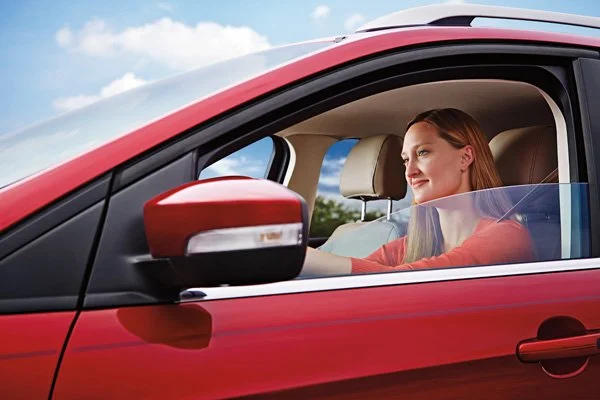 A woman with red hair driving a red car with a clear windshield, looking out with a smile against a partly cloudy sky.