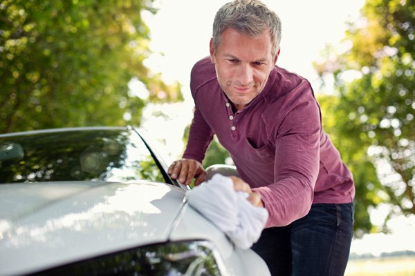 A man cleaning the hood of a silver car outdoors on a sunny day.