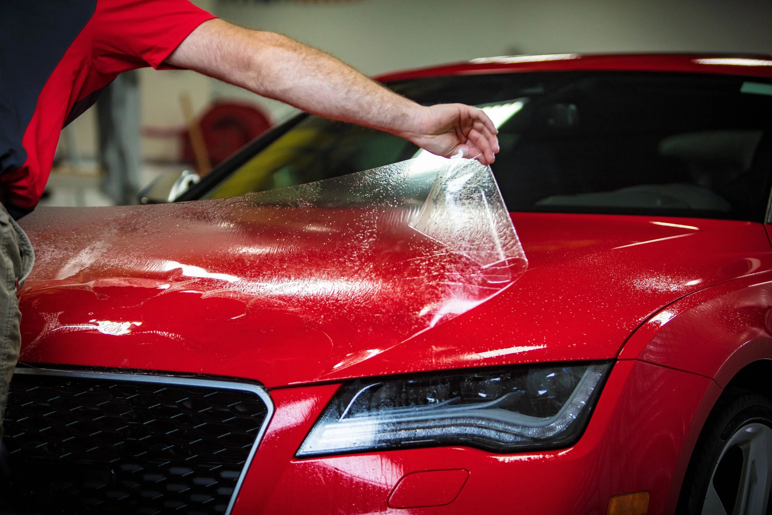 A person applying a clear protective film to the hood of a red sports car in a garage.