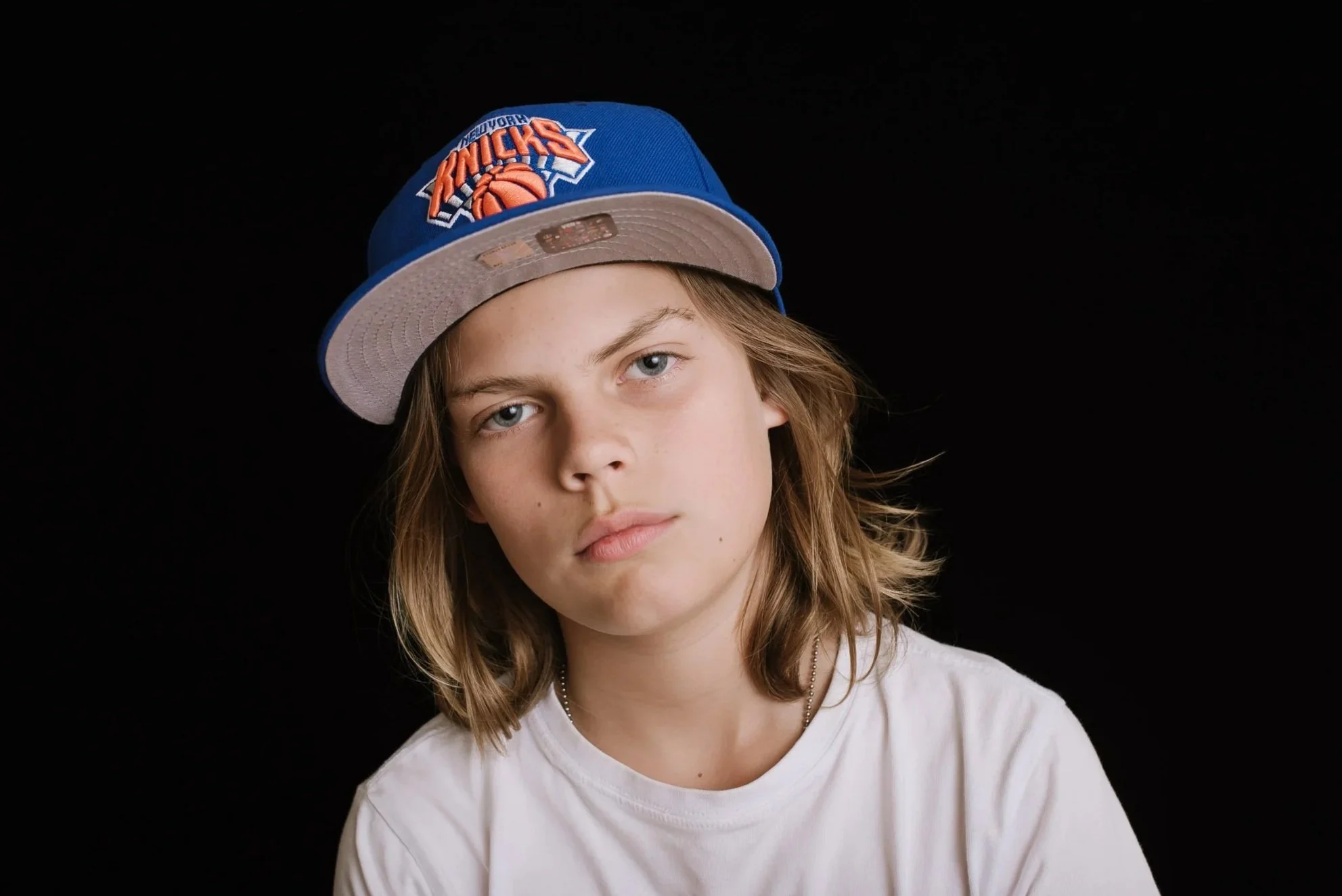 White boy in middle school wearing a baseball cap and a white shirt staring at the camera without smiling