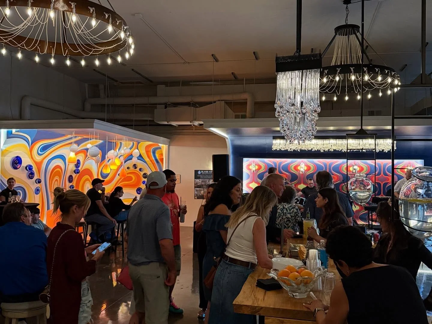 People gathered in a modern  event space decorated with colorful, abstract wall murals and hanging glass chandeliers.