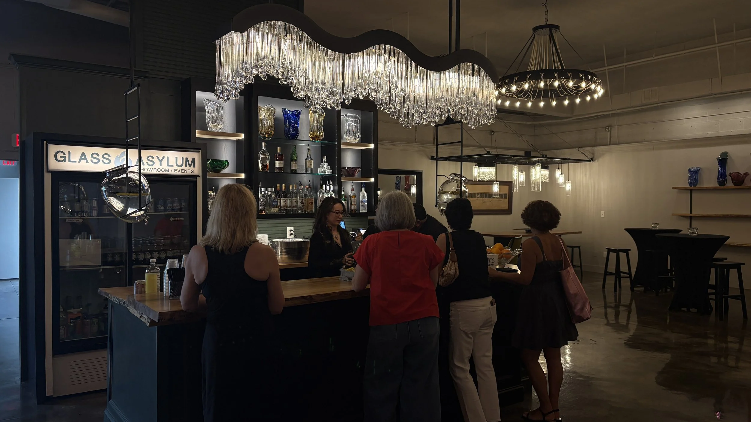 People gathered around a bar inside a dimly lit event space showroom with glassware and bottles on display, a fridge, and hanging chandeliers and wall décor.