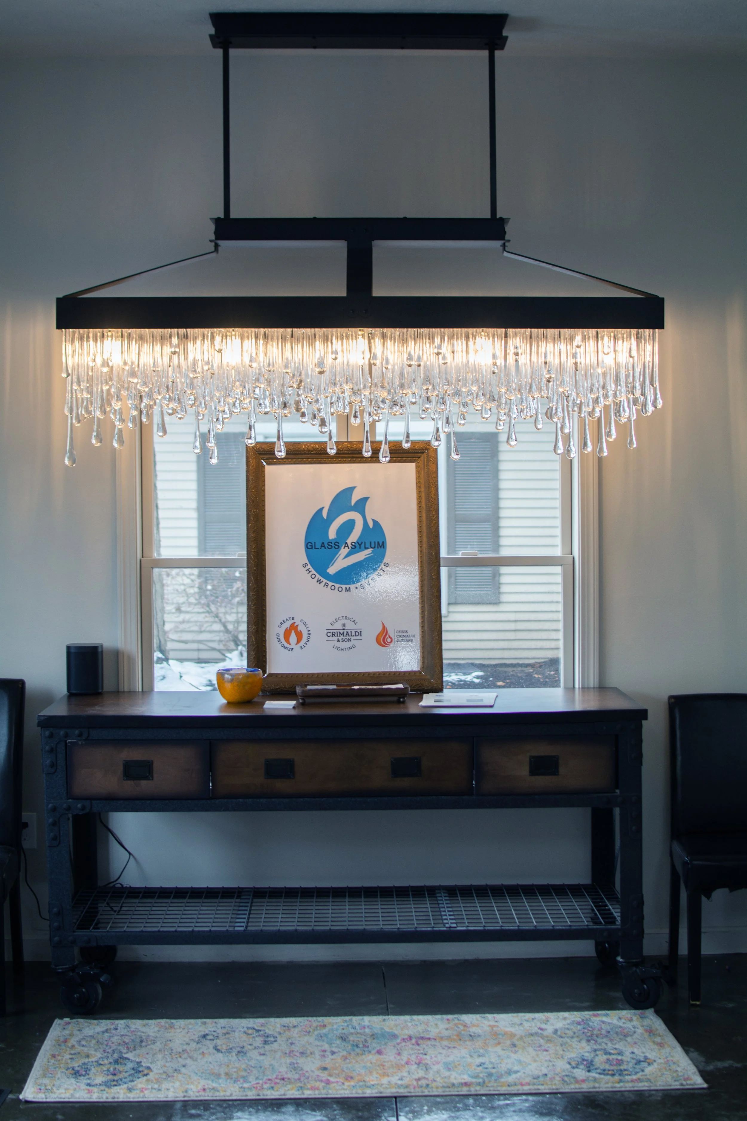 Chocolate-colored console table with a framed sign for Glass Asylum Showroom & Events, a small yellow bowl, and a notepad on top. A chandelier with hanging glass droplets hangs from the ceiling. Behind, a window with partially opened blinds shows a snow-covered landscape outside.