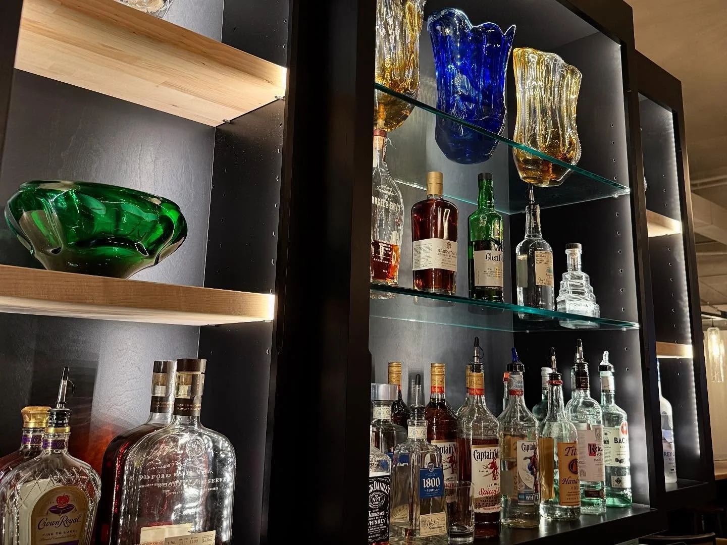 A display of various liquor bottles and colorful glassware inside a black display cabinet with glass shelves.