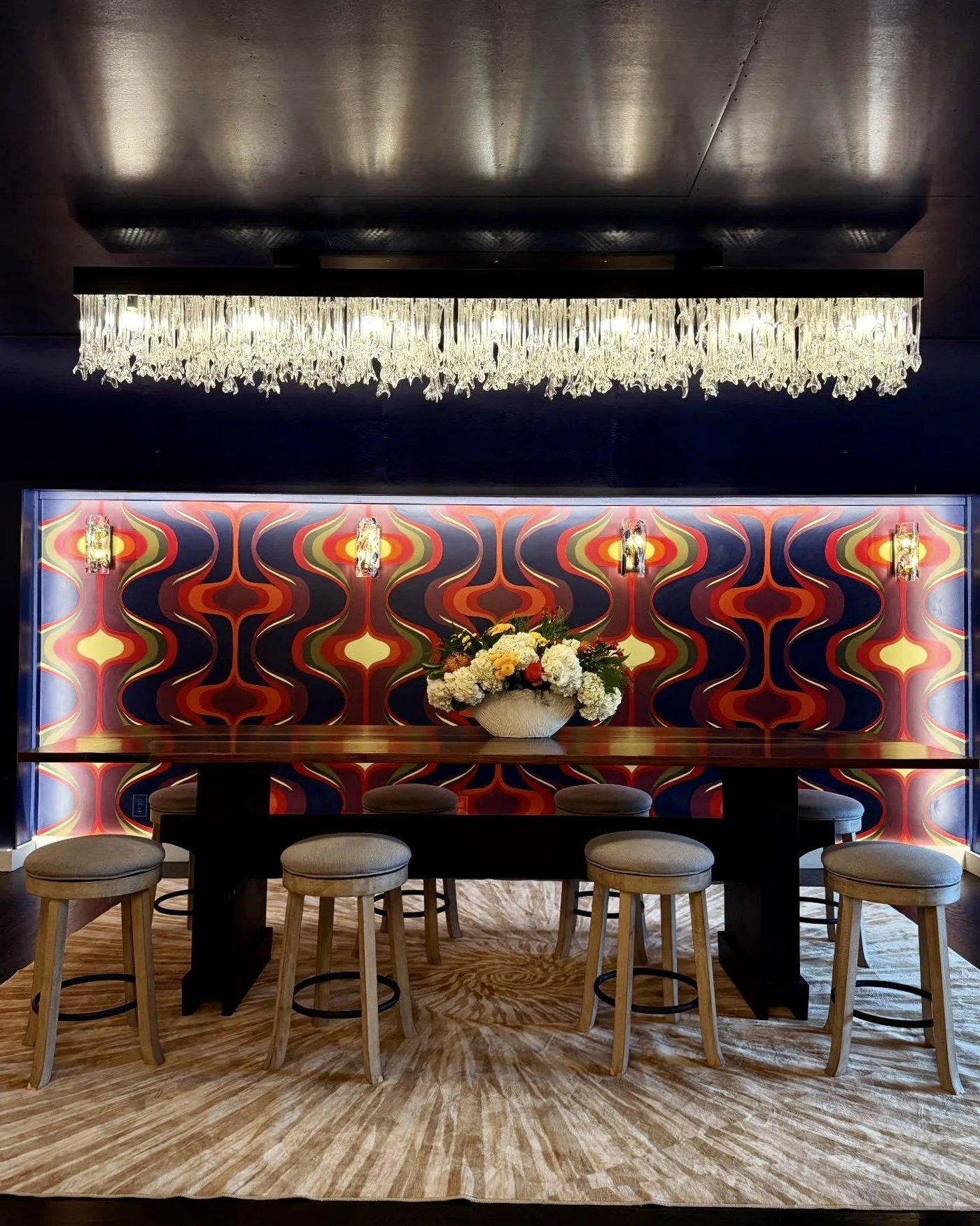 Elegant stage area in the venue with a long wooden table, six beige upholstered stools, a floral centerpiece, colorful retro-style patterned wallpaper, a crystal chandelier above, and a beige and brown area rug.