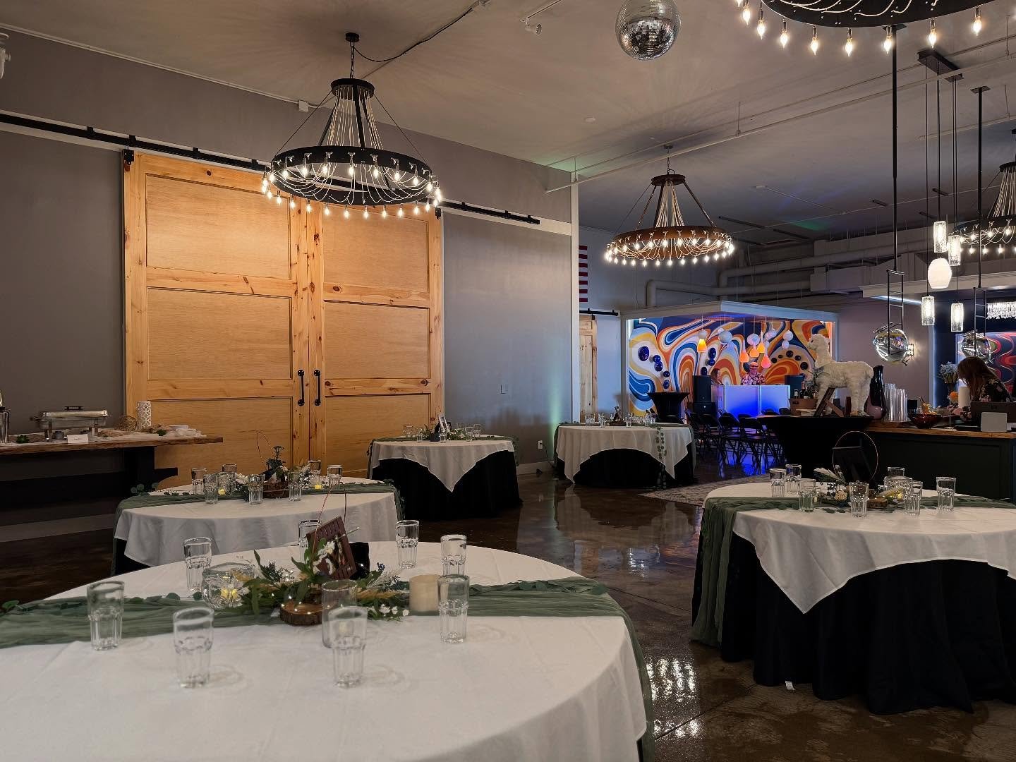 Indoor event space with decorated tables, custom overhead lights, a colorful abstract mural, and hanging chandeliers, prepared for a wedding celebration. 