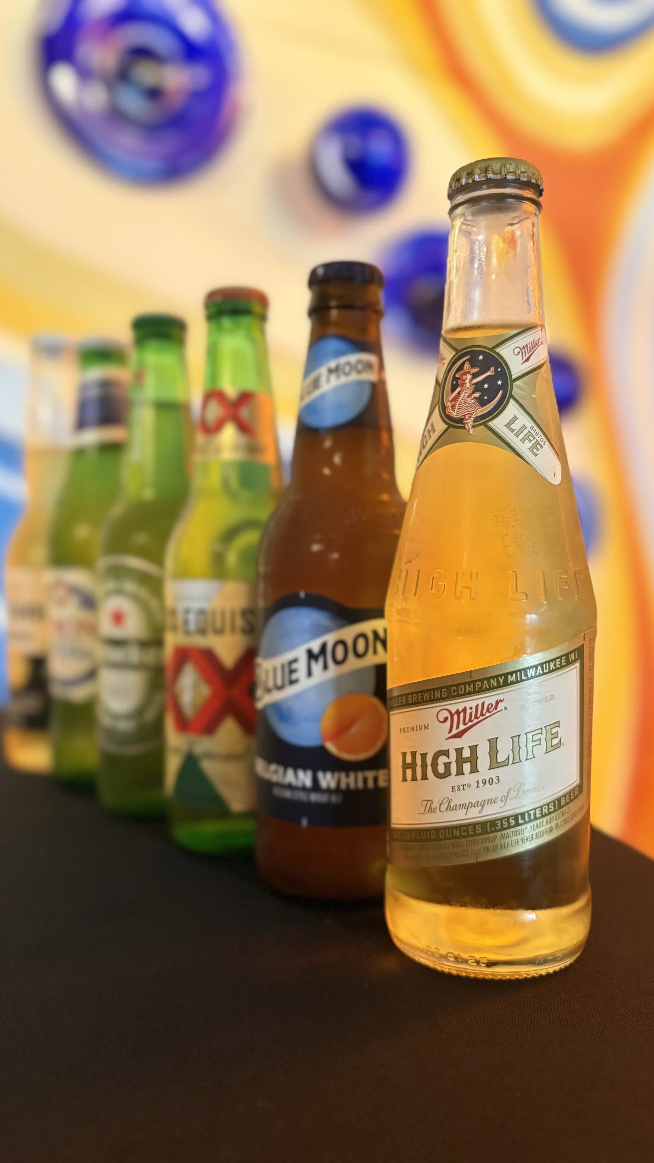 A lineup of various beer bottles including Miller High Life and Blue Moon, with a colorful abstract background.