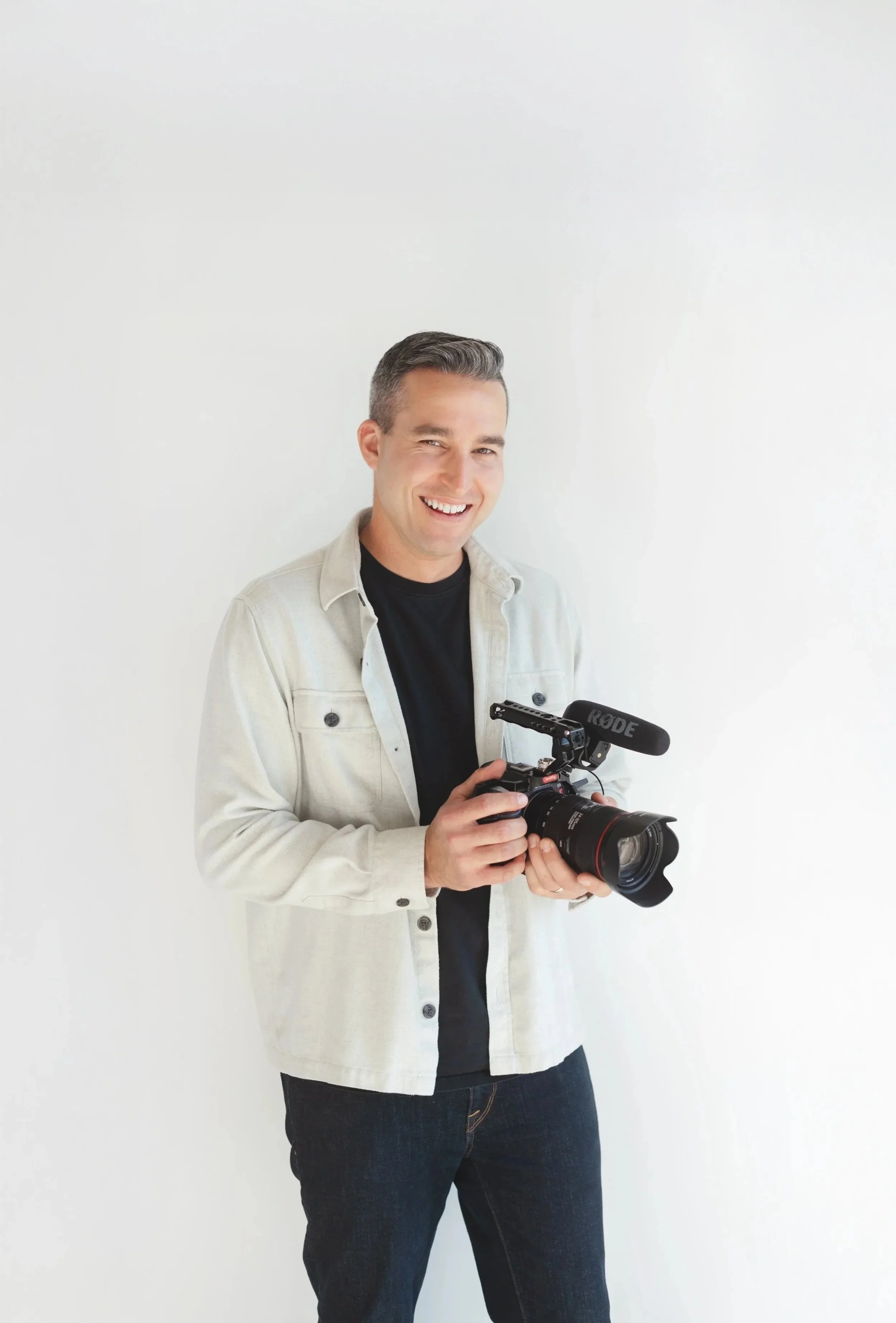 A man with short dark hair smiling while holding a professional camera with a Rode microphone attached, standing against a plain white wall.