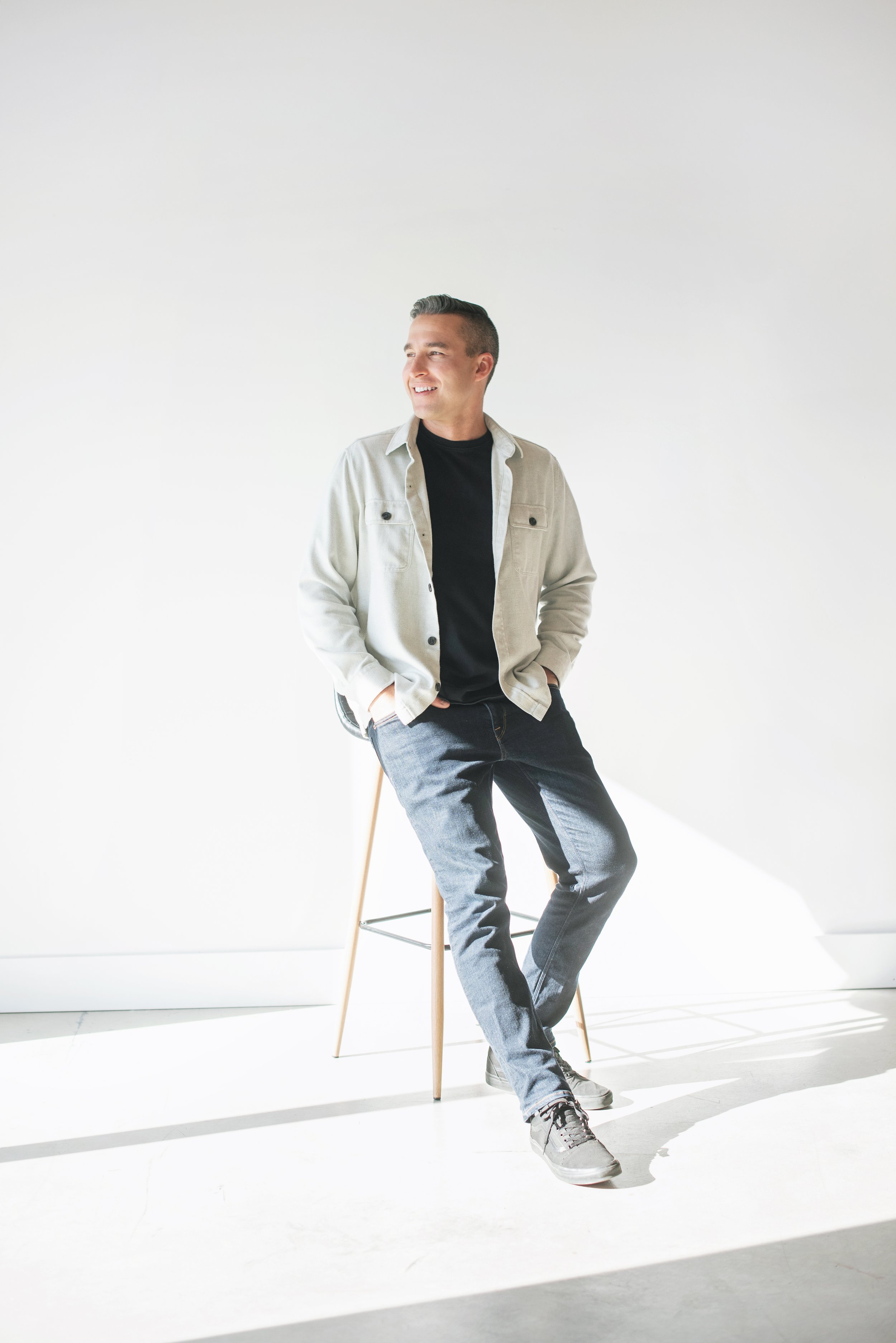 A man with dark hair styled upward, wearing a light-colored jacket over a black t-shirt, gray jeans, and sneakers, is standing in front of a plain white wall. He is smiling and looking to his left with one hand in his pocket and the other resting on his waist. He is sitting on a wooden chair with metal legs.