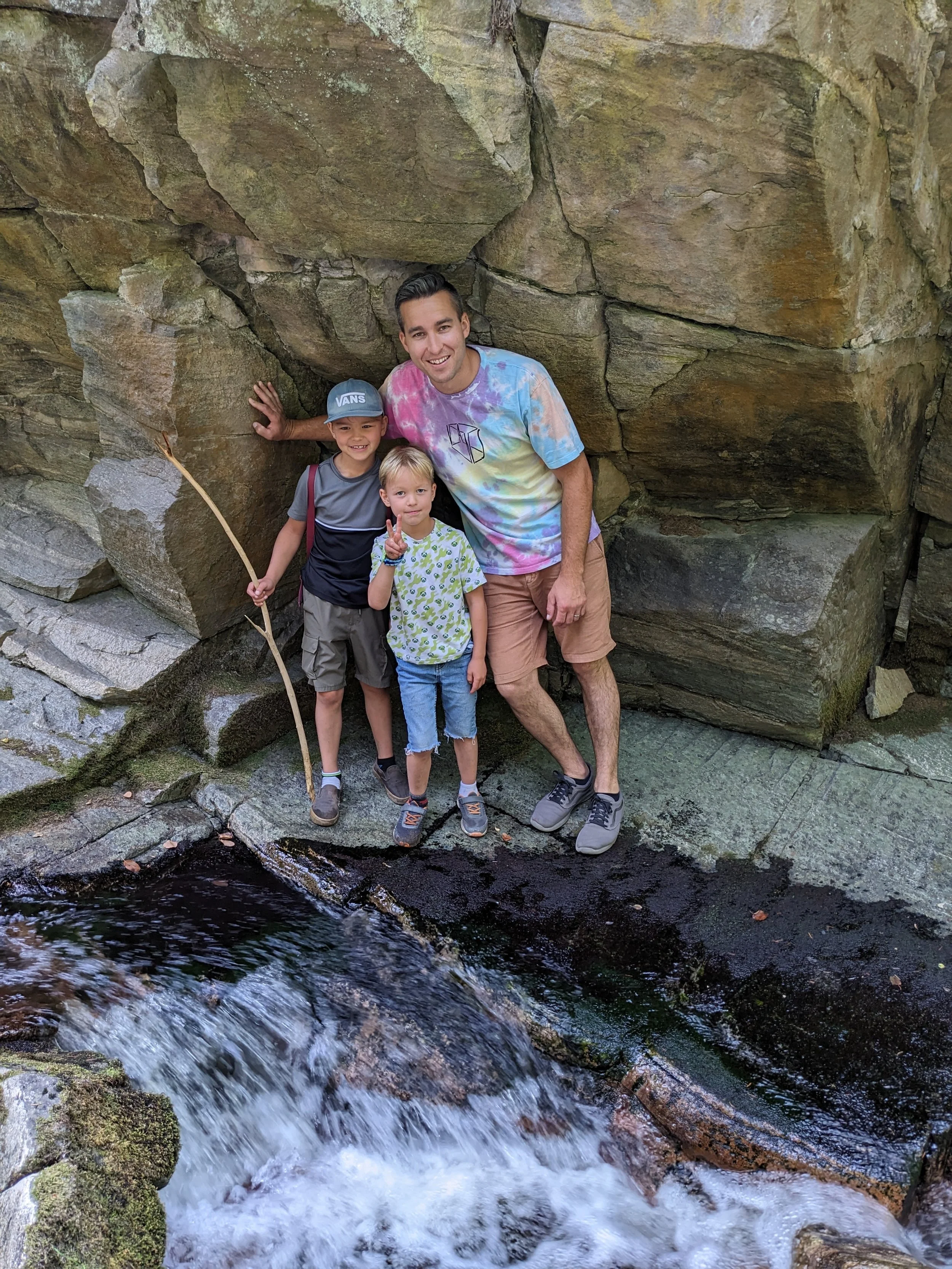 A man and two children standing on a rock ledge beside a small waterfall among large rocks and moss, smiling at the camera.
