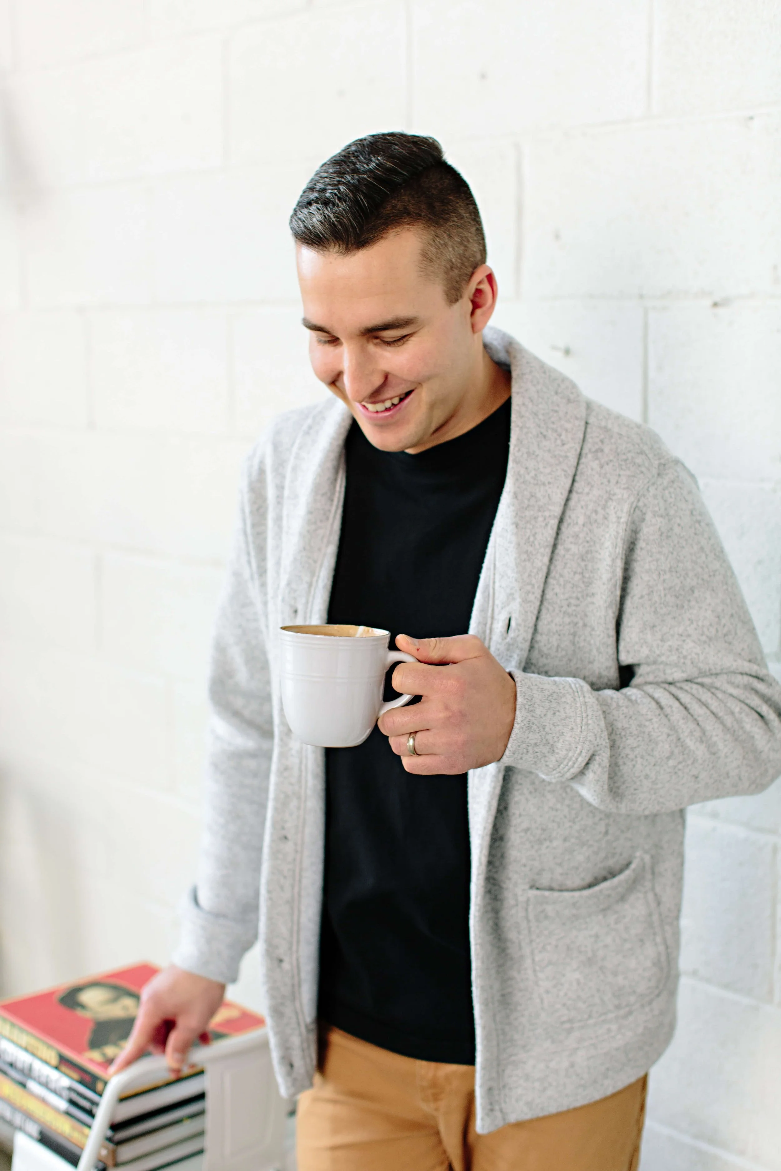 A man with dark hair styled in a slicked-back undercut wearing a grey hoodie, black t-shirt, and brown pants, holding a white coffee mug and smiling, standing in a room with white brick walls and a stack of books in the corner.