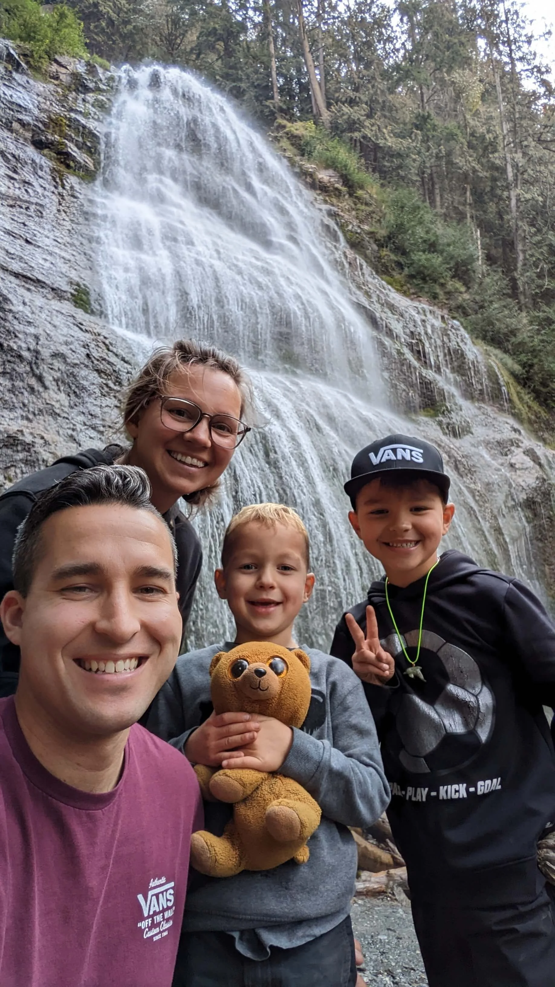 Four people taking a selfie in front of a waterfall surrounded by trees. They are smiling and appear happy. One child is holding a teddy bear. The group includes two adults and two children.