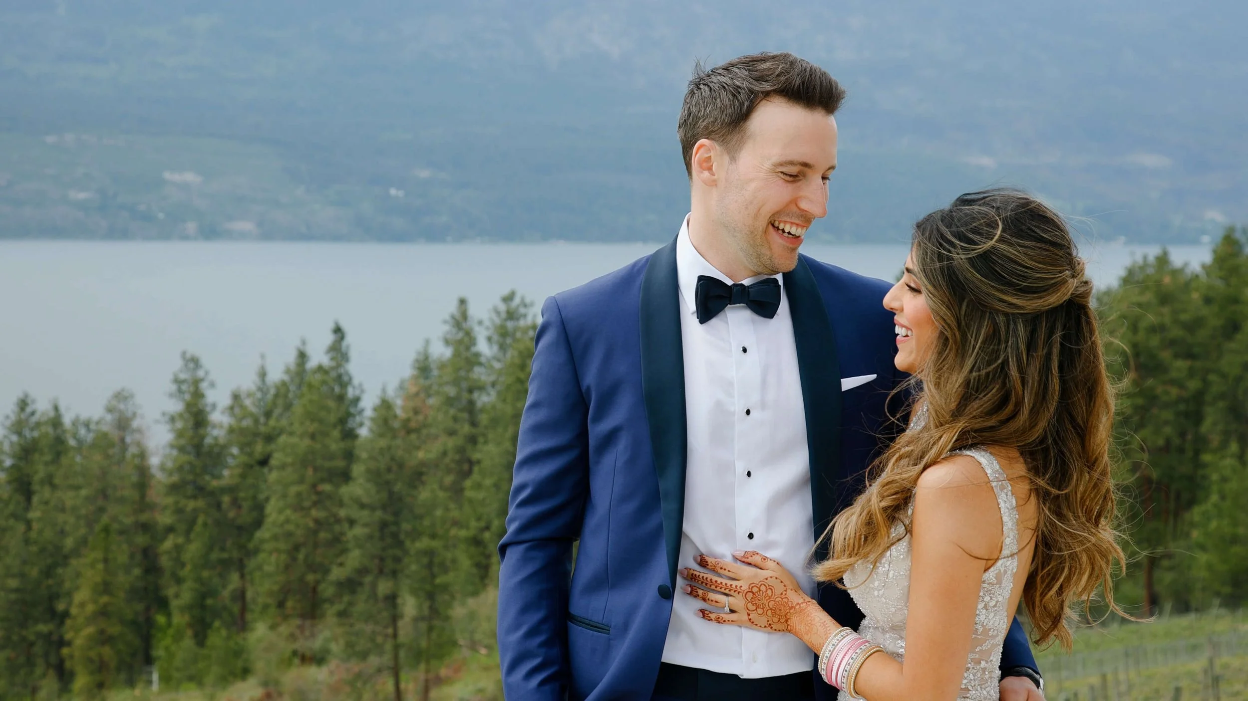 A newlywed couple smiling and gazing at each other outdoors with a lake and mountains in the background.