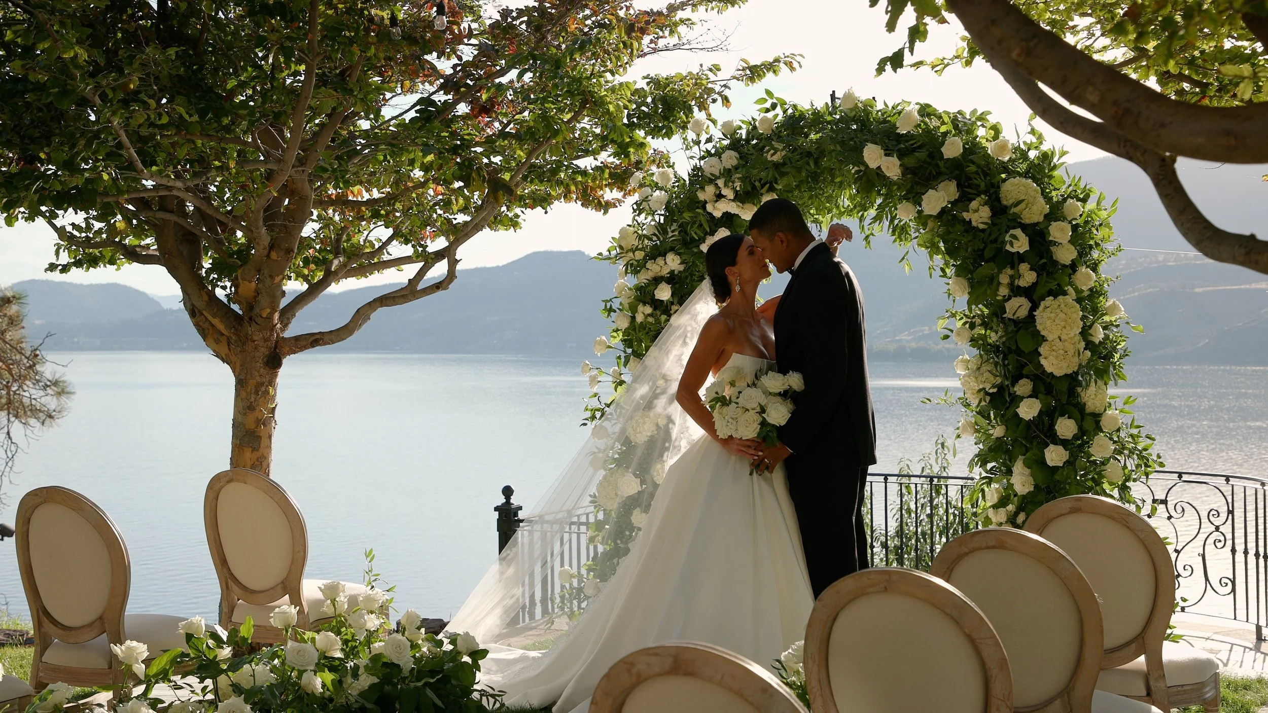 Bride and groom sharing a kiss during their outdoor wedding ceremony by a lake, framed by a floral arch with white roses and green foliage, with mountains in the background.