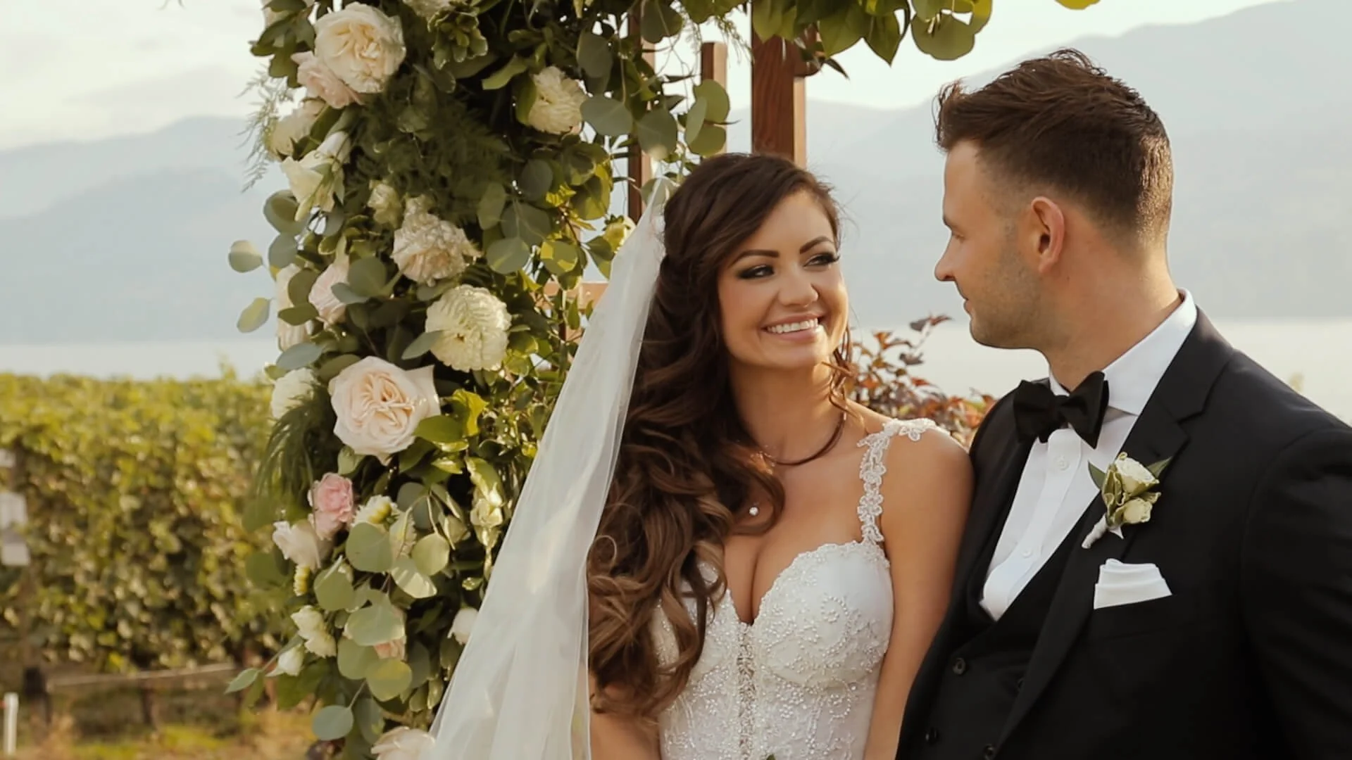 A bride and groom standing outdoors at their wedding ceremony, smiling at each other, with a floral arch decorated with white and pale pink flowers and green foliage behind them.
