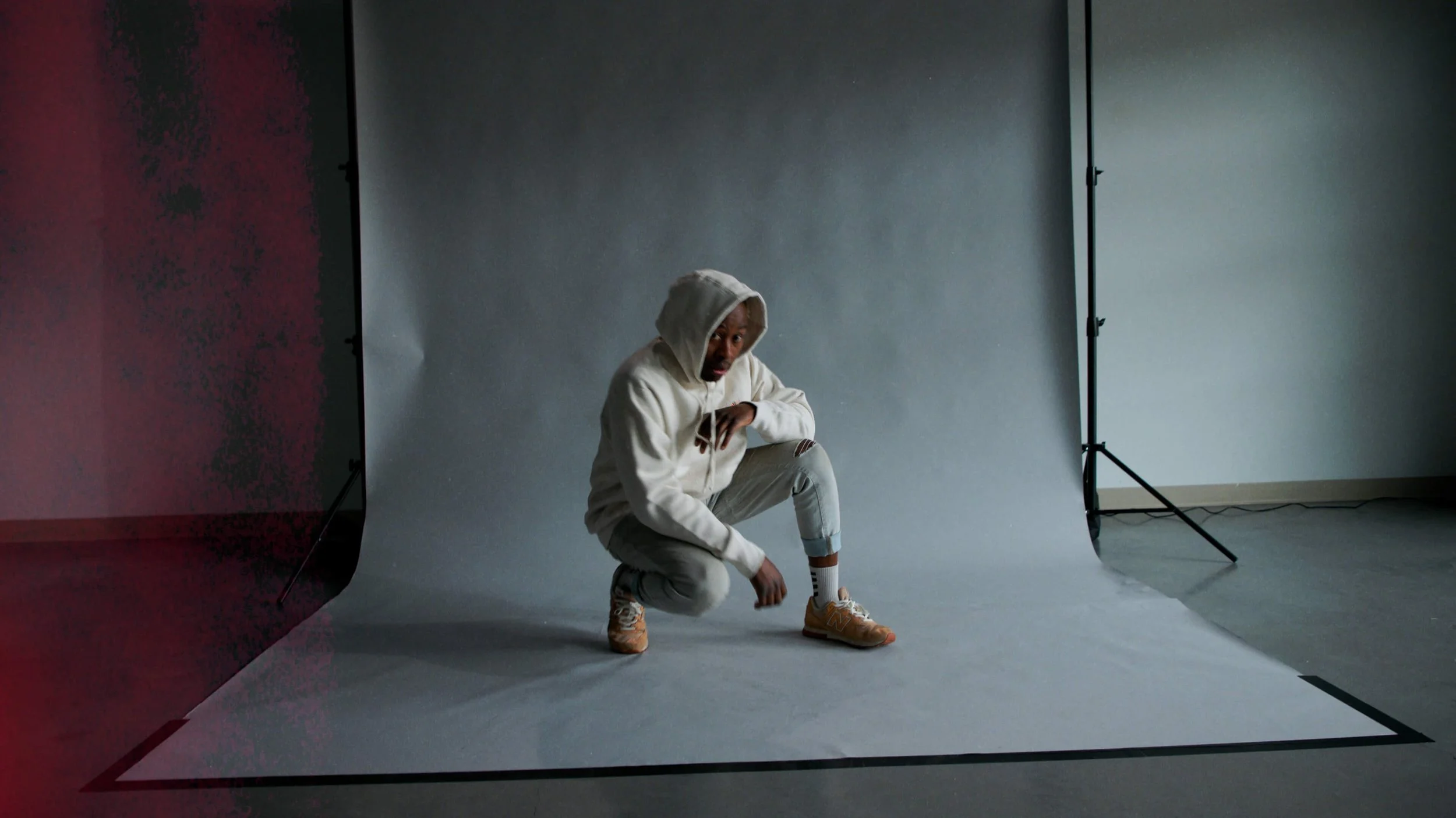 A person squatting on the floor in a photography studio with a plain gray backdrop and soft lighting, wearing a hooded sweatshirt, distressed jeans, and sneakers.