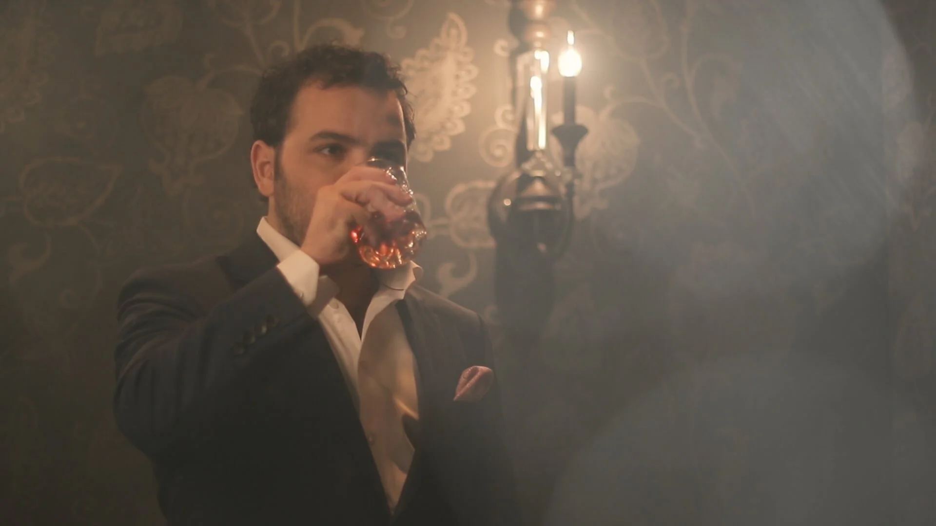 A man in a suit and white shirt drinks from a glass of whiskey in a dimly lit room with dark patterned wallpaper and a wall-mounted lamp.