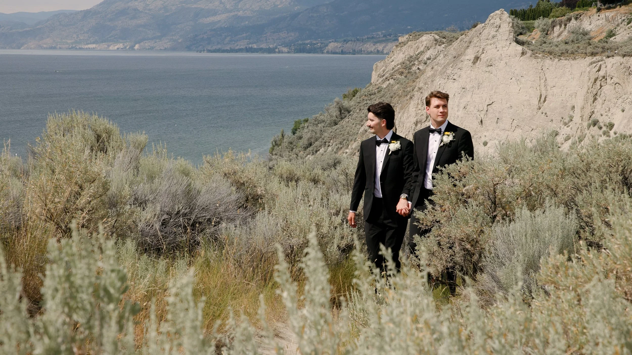 Two men in tuxedos holding hands while walking in a natural coastal landscape with shrubs, cliffs, and a body of water in the background.
