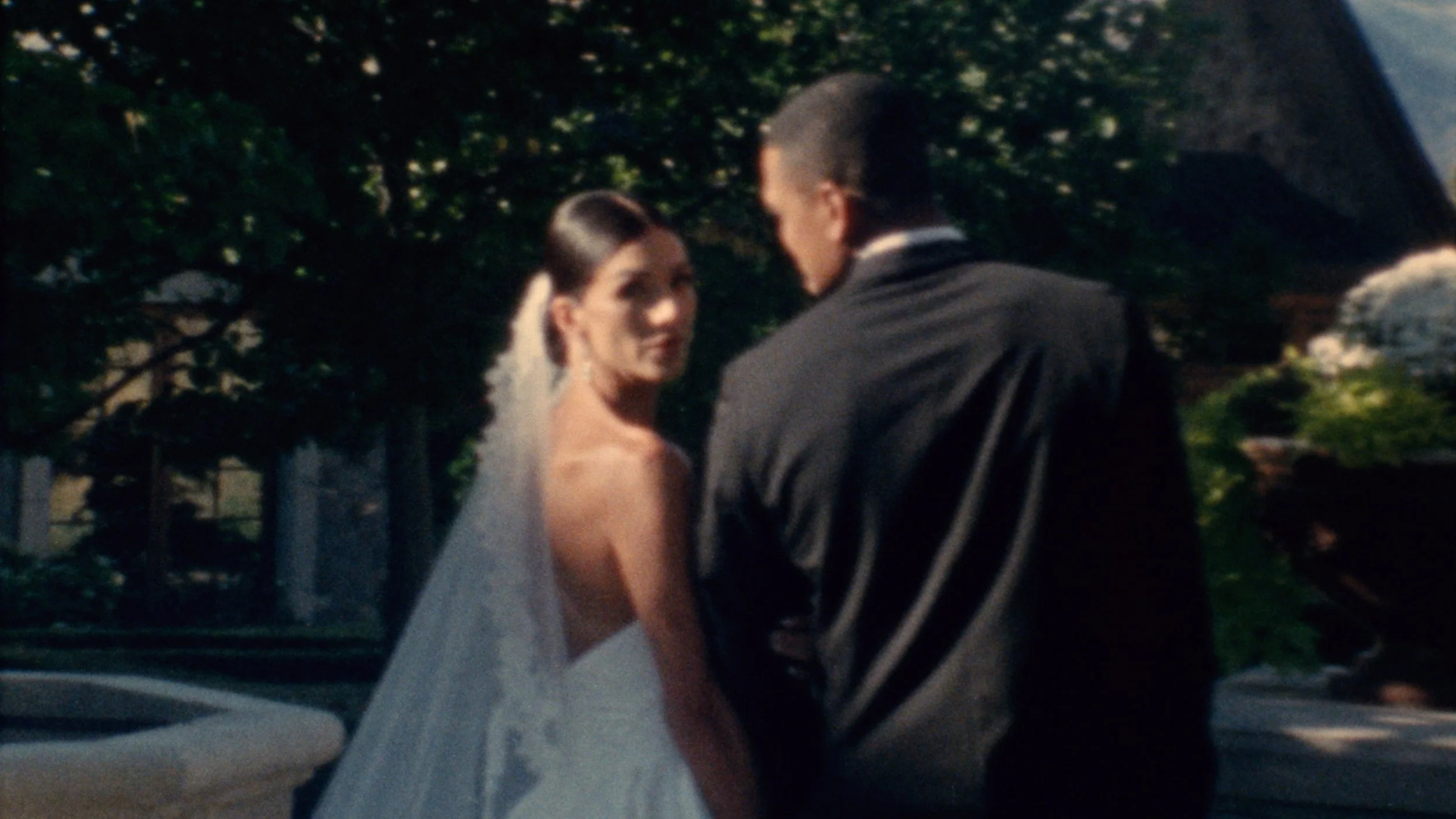 A bride with dark hair styled in an updo and wearing a white wedding gown and veil, looking over her shoulder at a groom with short dark hair, dressed in a dark tuxedo, outdoors with trees and greenery in the background.