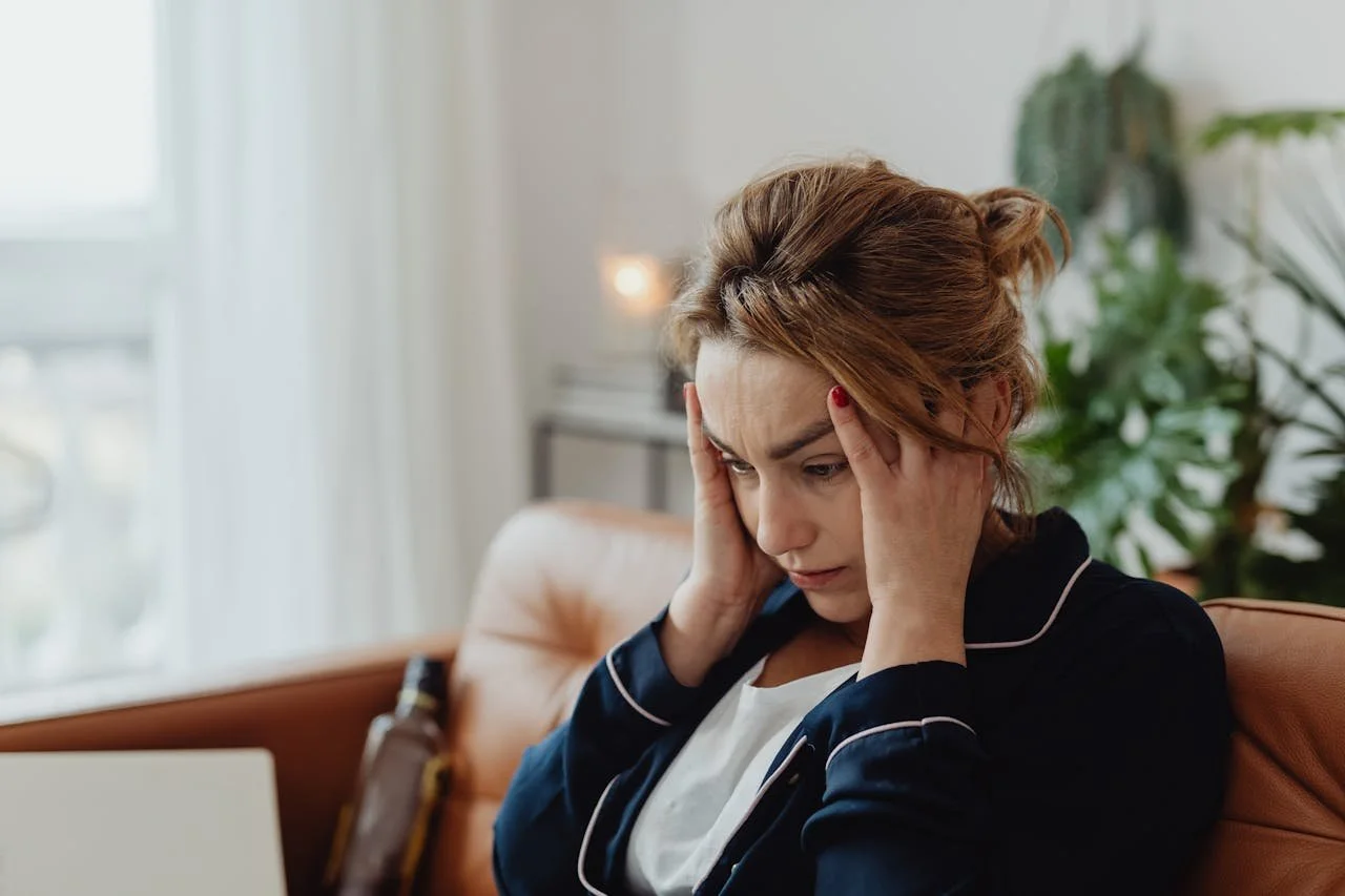 Woman feeling overwhelmed and stressed, holding her head while sitting on a couch—representing holiday perfectionism, emotional overwhelm, and anxiety experienced by high-achieving women.
