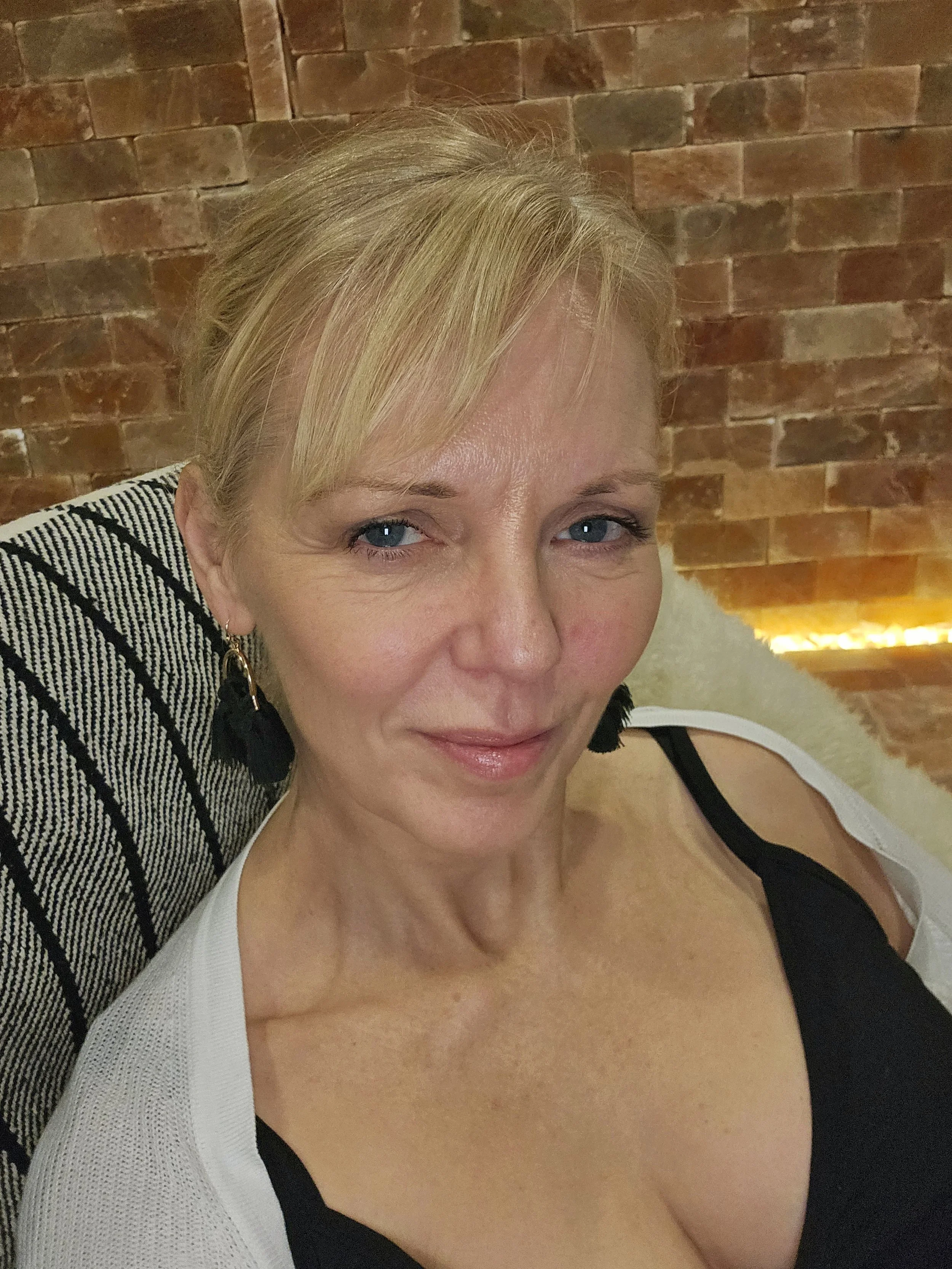 Close-up of a woman with blonde hair, wearing a black top and cardigan, standing in front of a brick wall with warm lighting.