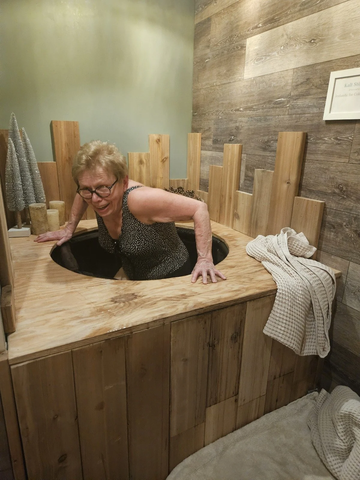 An older woman with glasses and short curly hair smiling as she leans out of a wooden hot tub or spa with her arms resting on the edge, surrounded by wooden walls and some decorative miniature trees.
