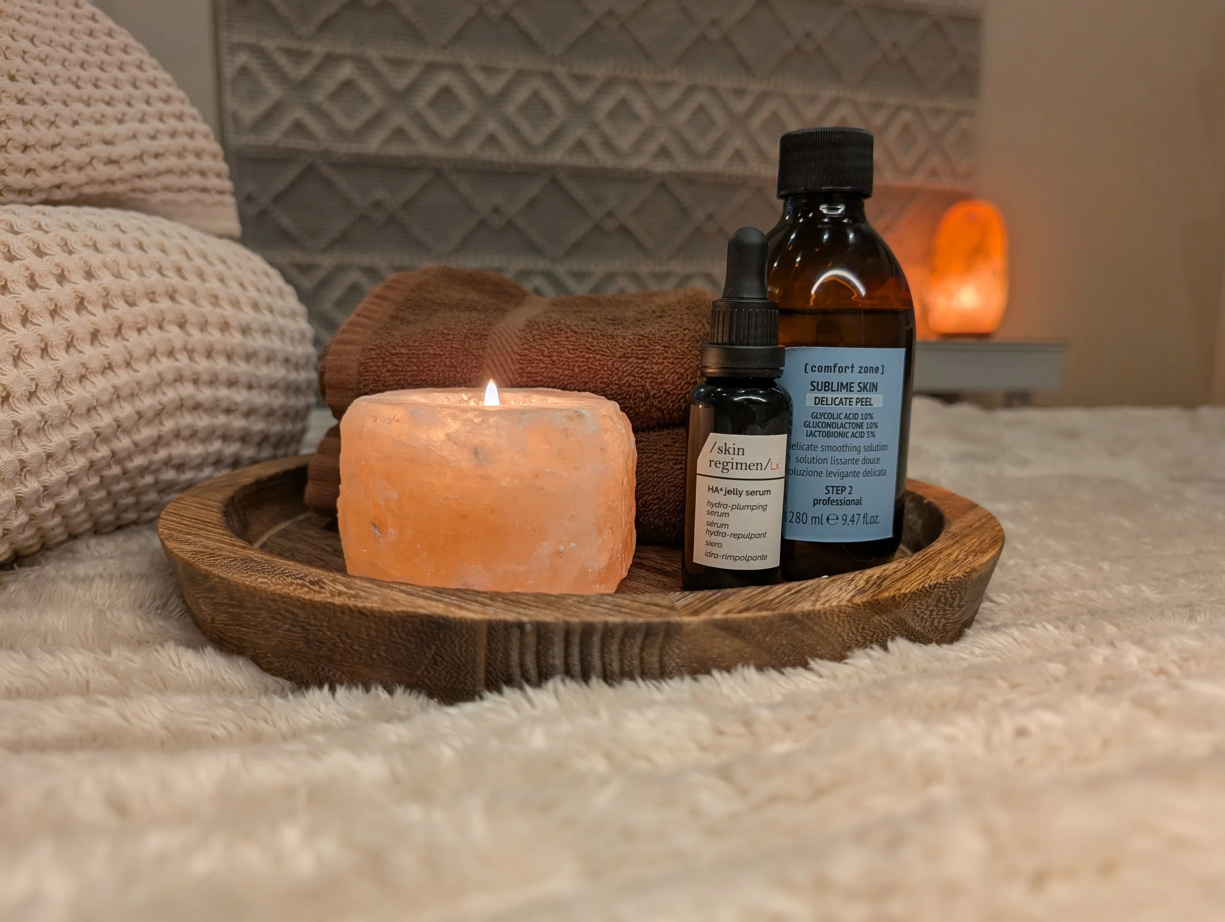 A wooden tray with [comfort zone] products and a glowing Himalayan salt votive on a blanket beside cotton waffle weave towels with a rug wall hanging and a Himalayan salt lamp in the background.