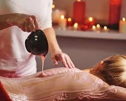 An aesthetician pouring cream from a treatment cup on the back of a female client who is lying comfortably with candles in the background.