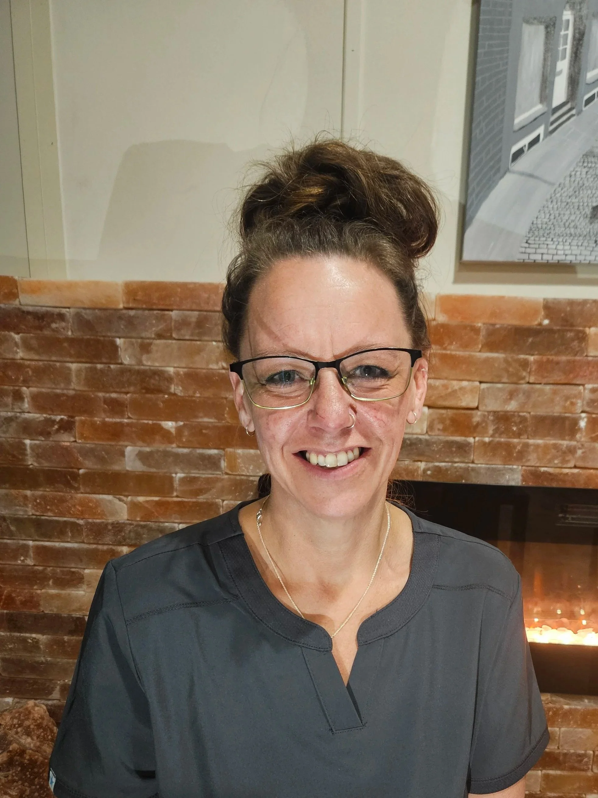 Smiling woman wearing glasses and a dark top, with a brick wall in the background.