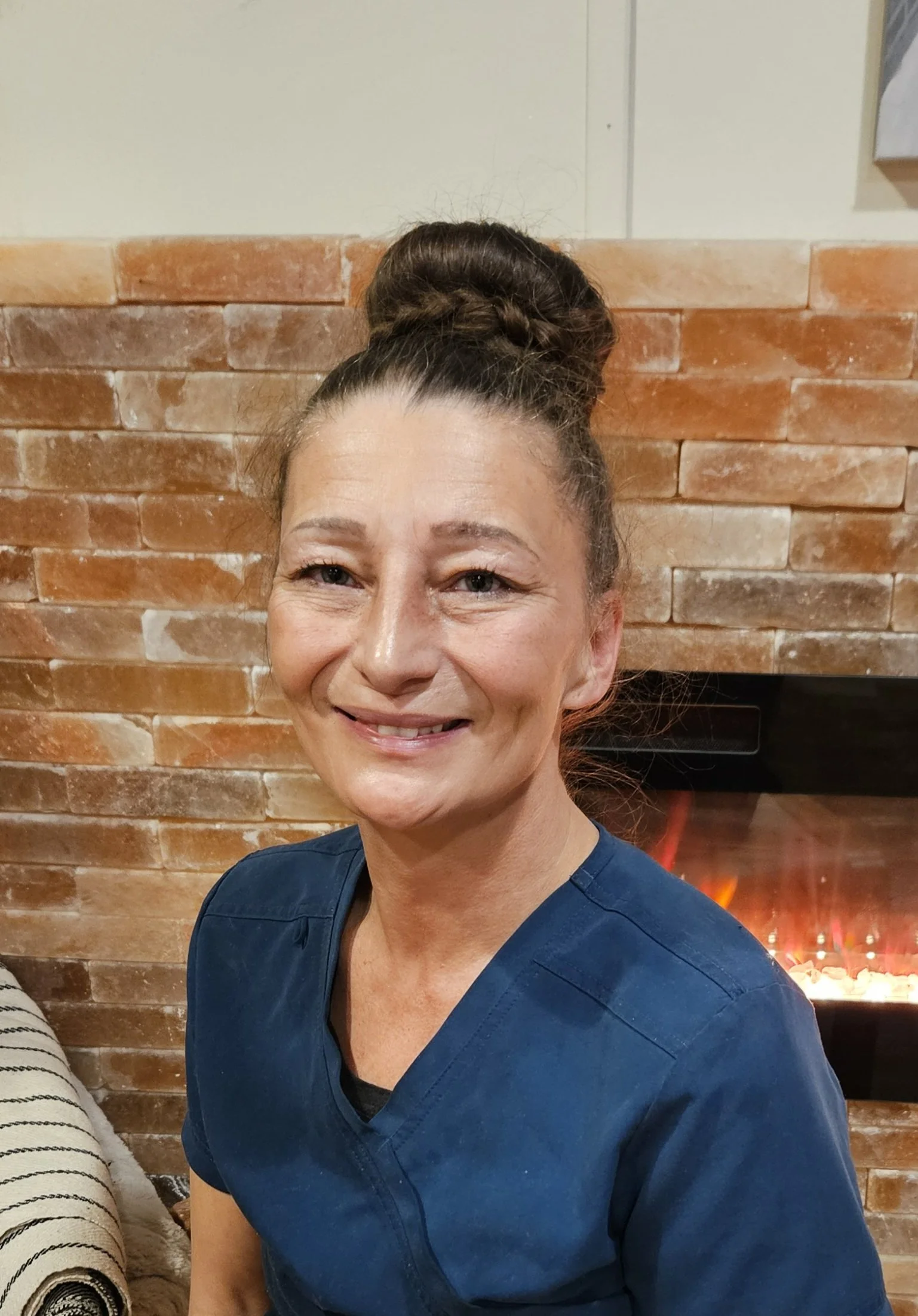 Woman with dark hair tied up, smiling, wearing navy blue scrubs, standing in front of a brick wall with a fireplace.