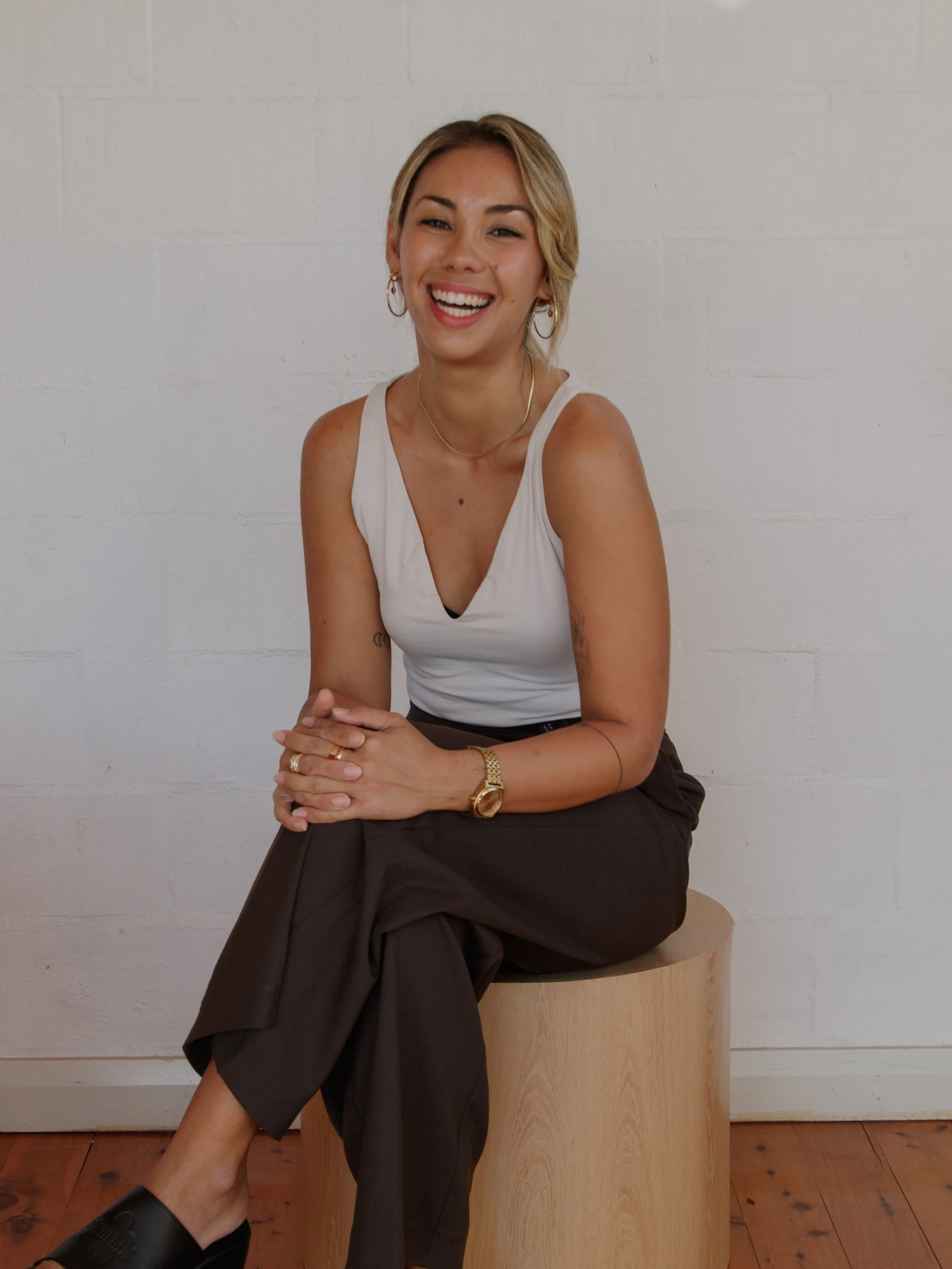 A woman with blonde hair, wearing a white sleeveless top, dark pants, and black slippers, is sitting on a wooden block against a white wall, smiling with her hands clasped.