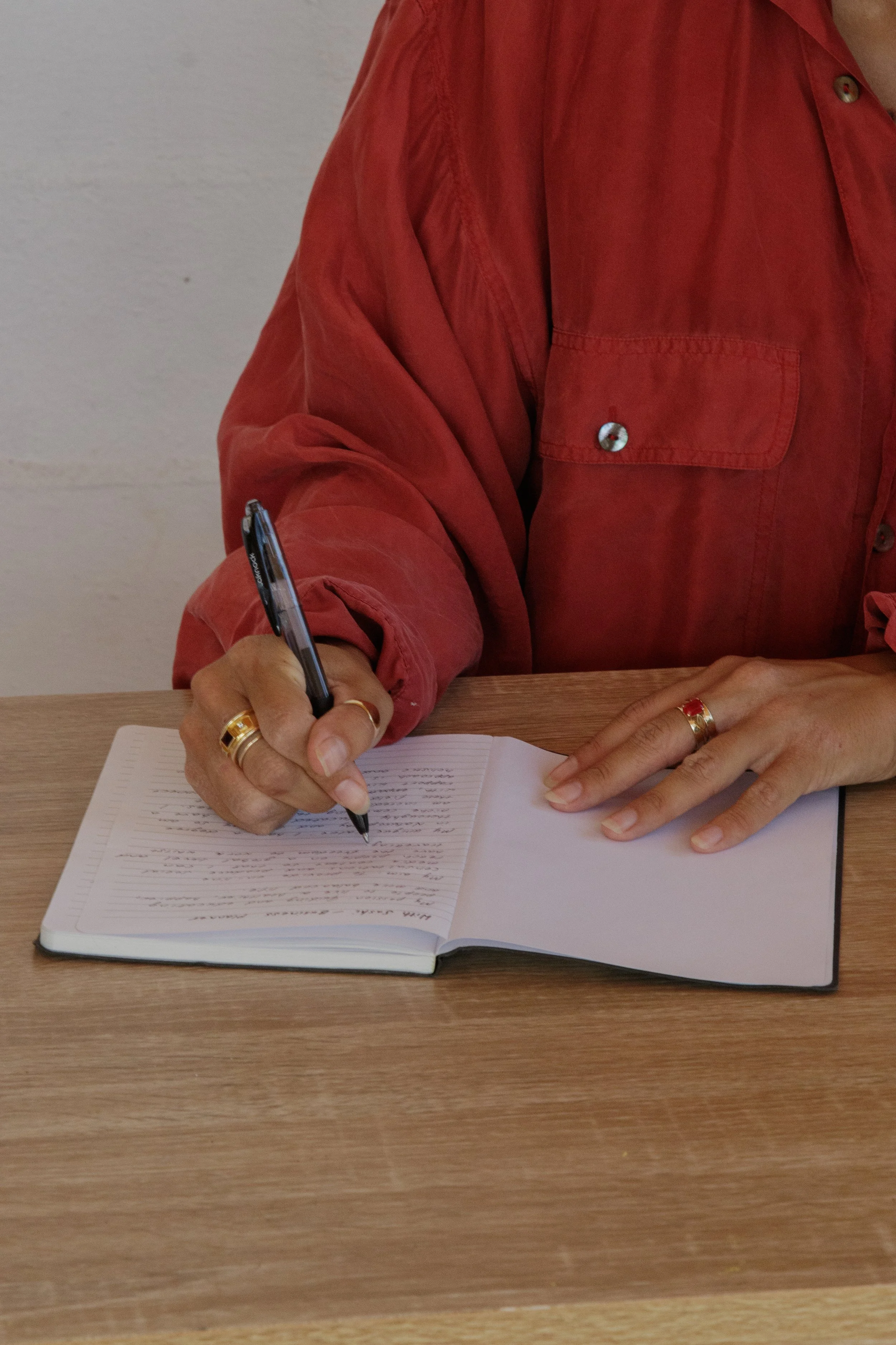 Person wearing a red shirt writing in a notebook with a black pen on a wooden table.
