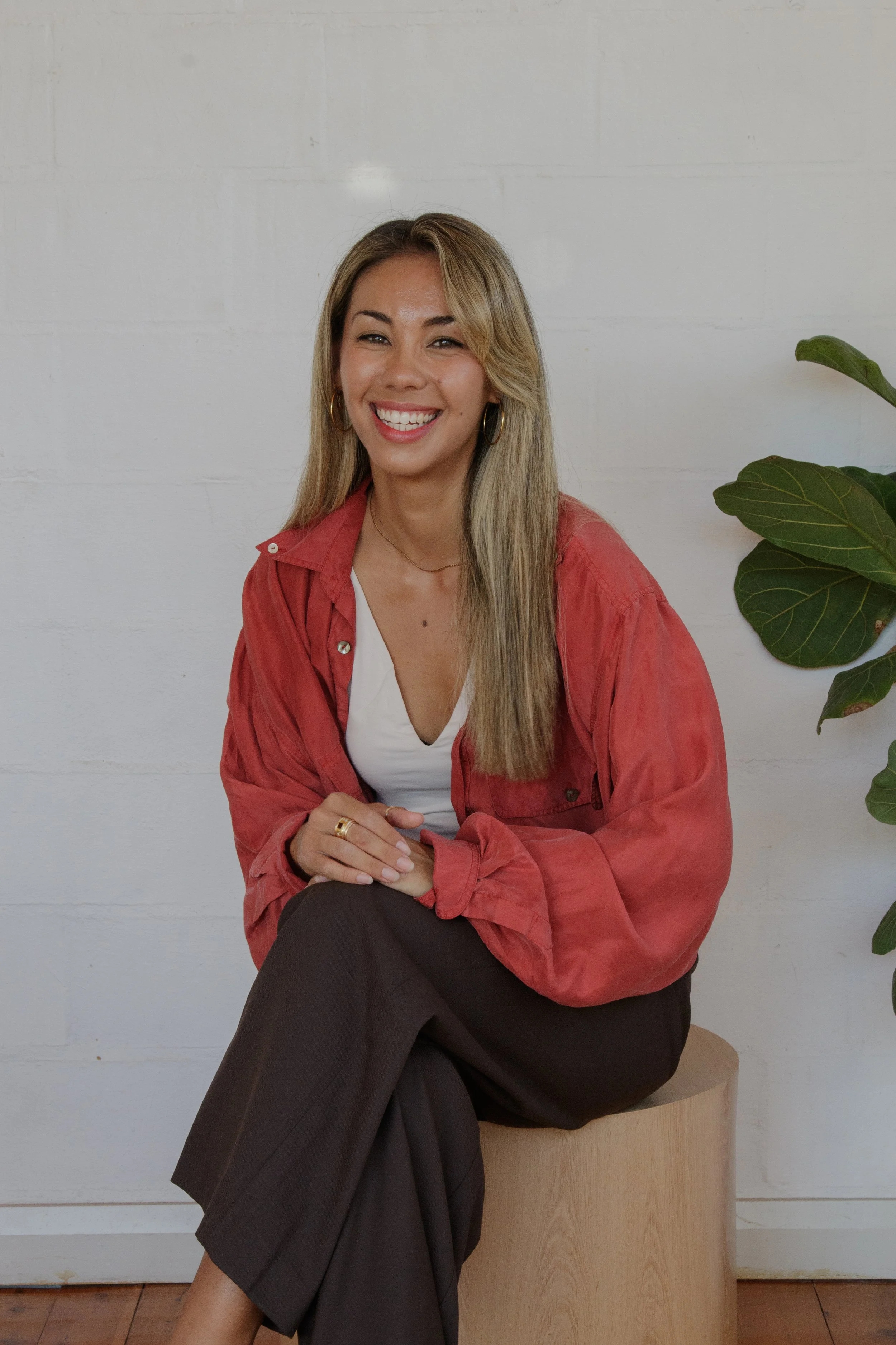 A woman with long blonde hair sitting on a wooden stool, wearing a red blazer over a white top, smiling, with green leafy plant nearby, in front of a white wall.