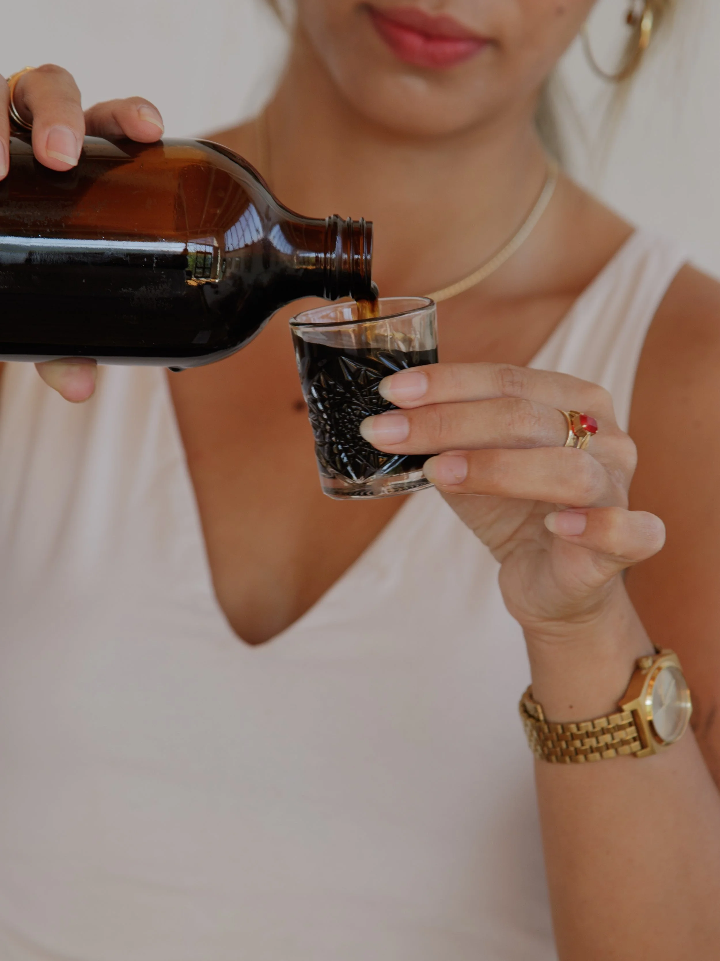 A woman pours a dark beverage from a brown glass bottle into a decorative glass mug, wearing a white sleeveless top, gold jewelry, and a gold watch.