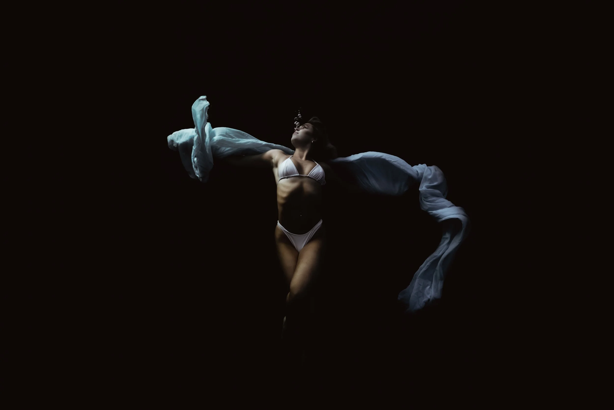 Person underwater with long hair, wearing a dark dress, against a purple background and red light.