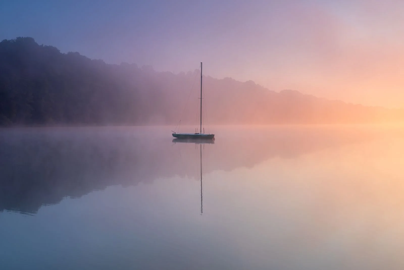 A sailboat floating on a calm lake surrounded by fog, with hills in the background during sunrise or sunset.