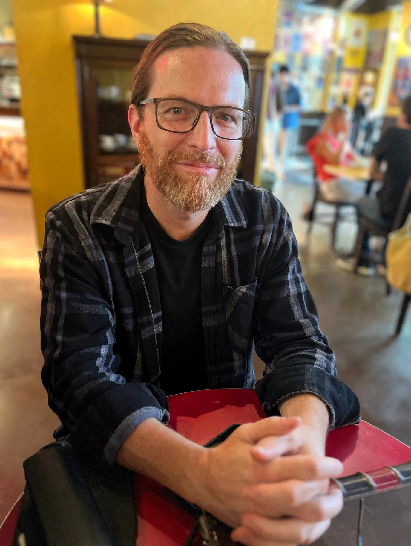 A man with glasses and a beard sitting at a table in a cafe or restaurant, with colorful walls and other patrons in the background.