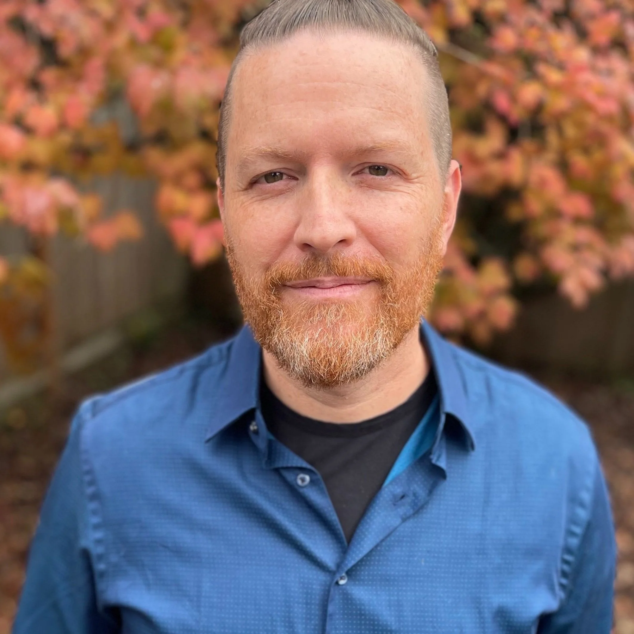 Close-up of a man's face with a red beard and short hair, wearing a blue button-up shirt, standing outdoors with blurred autumn-colored leaves in the background.