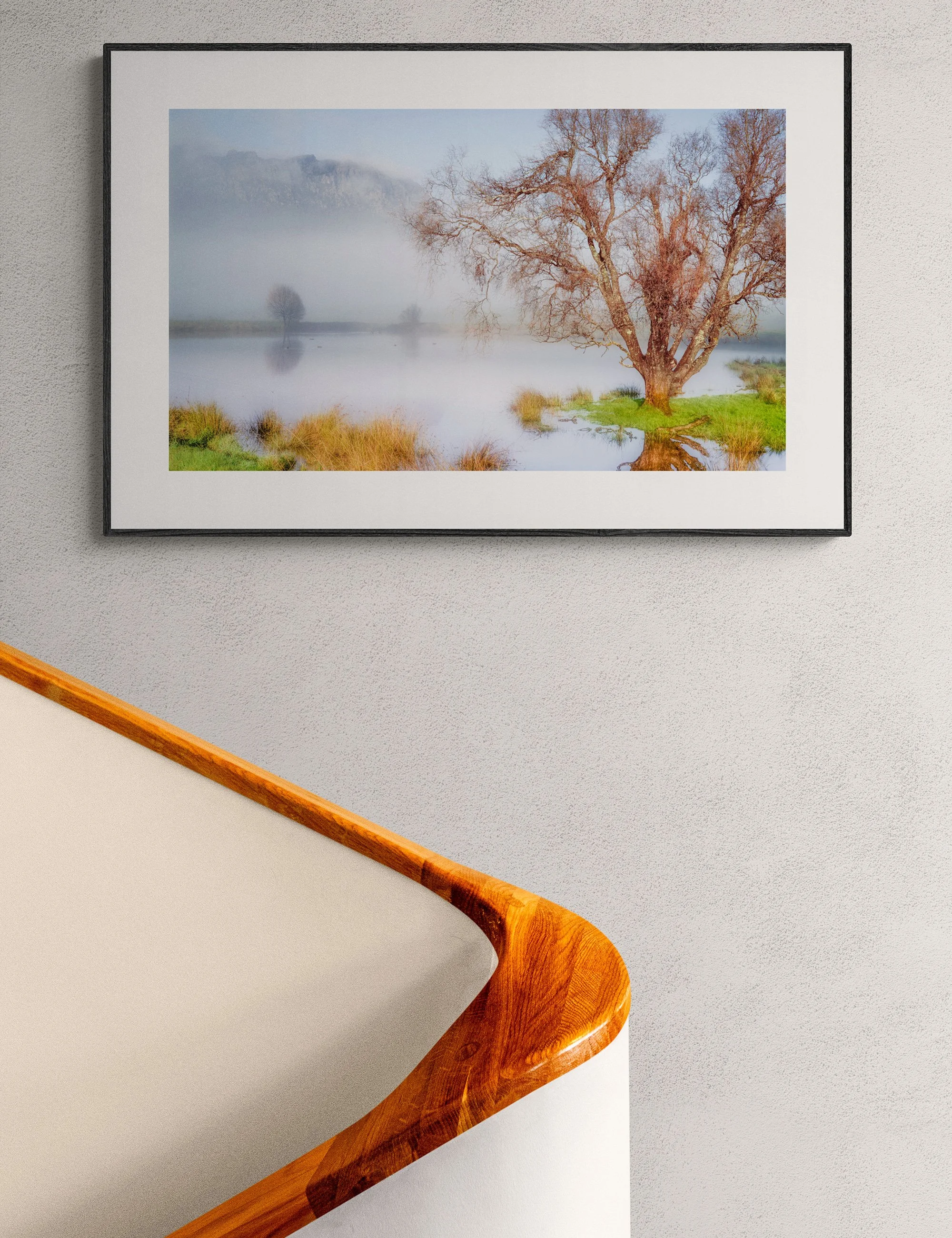 “Winter Tranquility” framed landscape print by Ian J. Muirhead Arts above a curved timber staircase in a minimalist interior. Misty trees and soft reflections create a calm rural atmosphere.