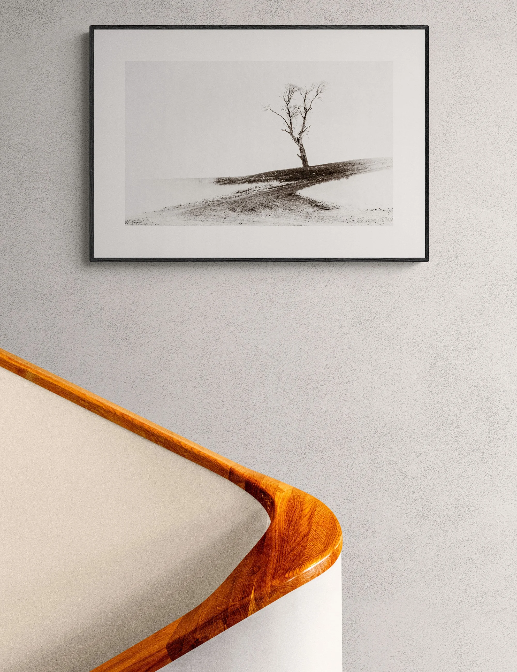 “A Harsh Land” framed minimalist landscape print by Ian J. Muirhead Arts above a curved timber staircase in a minimalist interior. A lone tree and sweeping tracks create a stark rural scene.