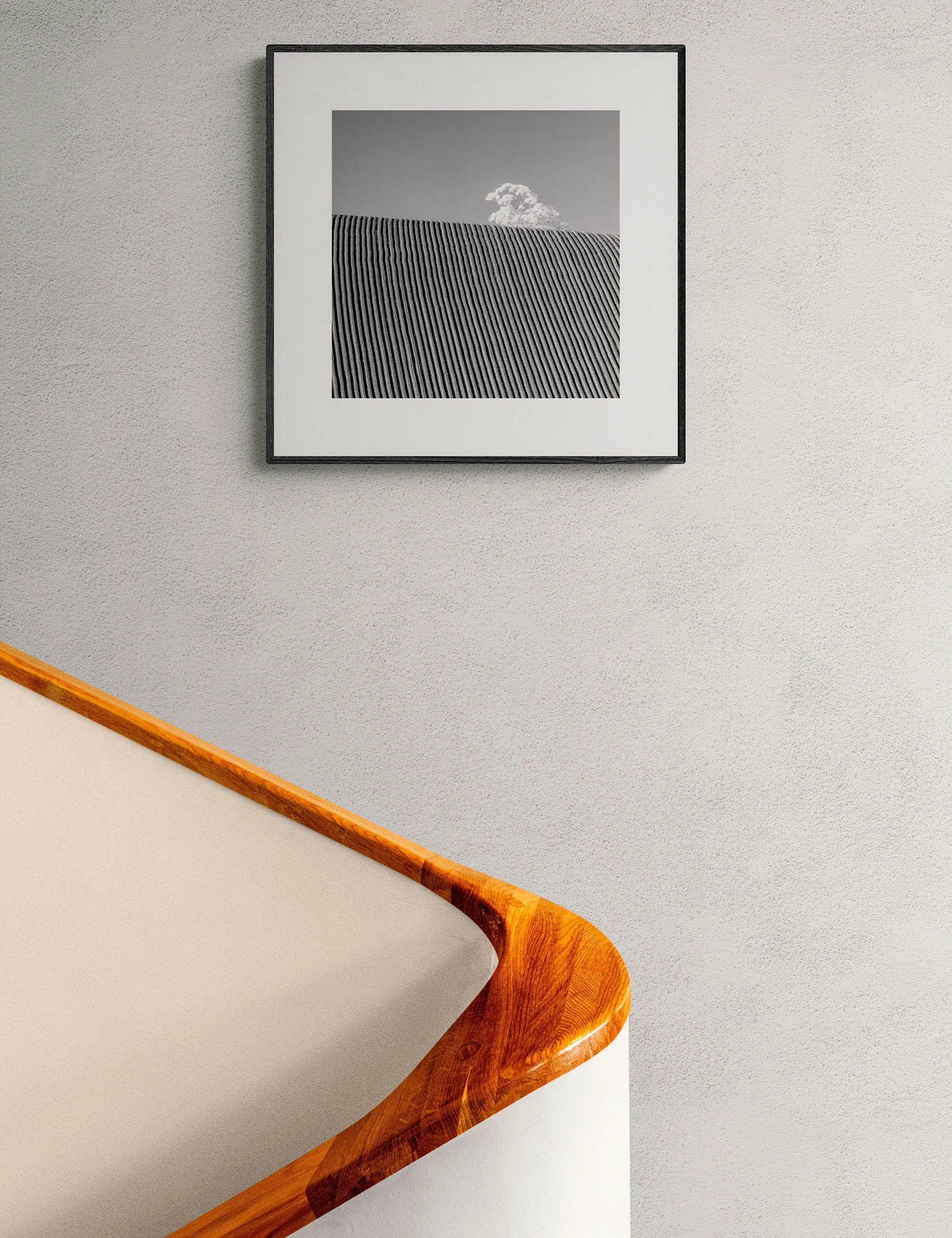 “Corduroy Country” framed monochrome landscape print by Ian J. Muirhead Arts above a curved timber staircase in a minimalist interior. Rhythmic field lines and a lone cloud create a minimalist scene.